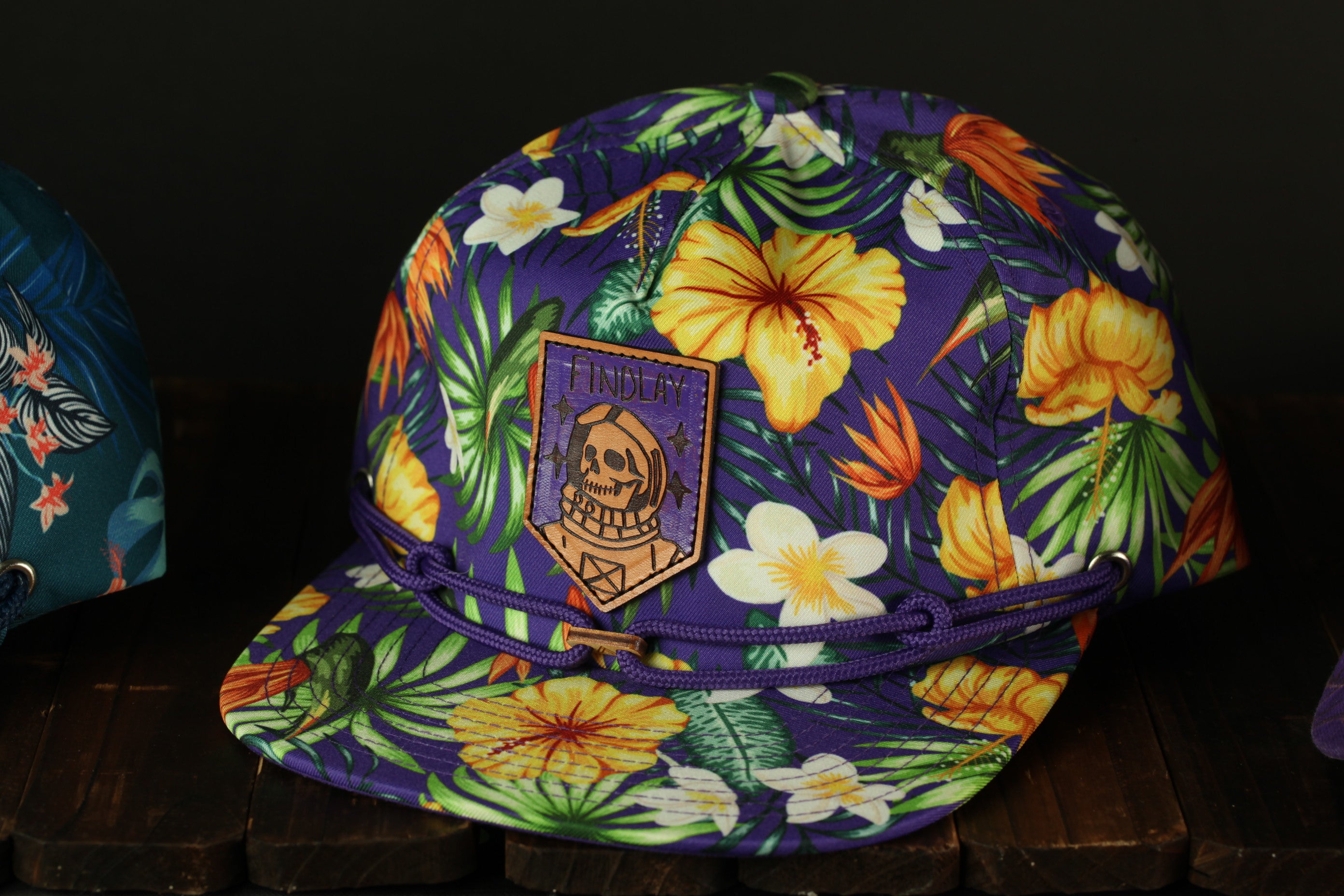 A colorful floral print baseball cap with a skull patch on the front, displayed on a wooden surface.