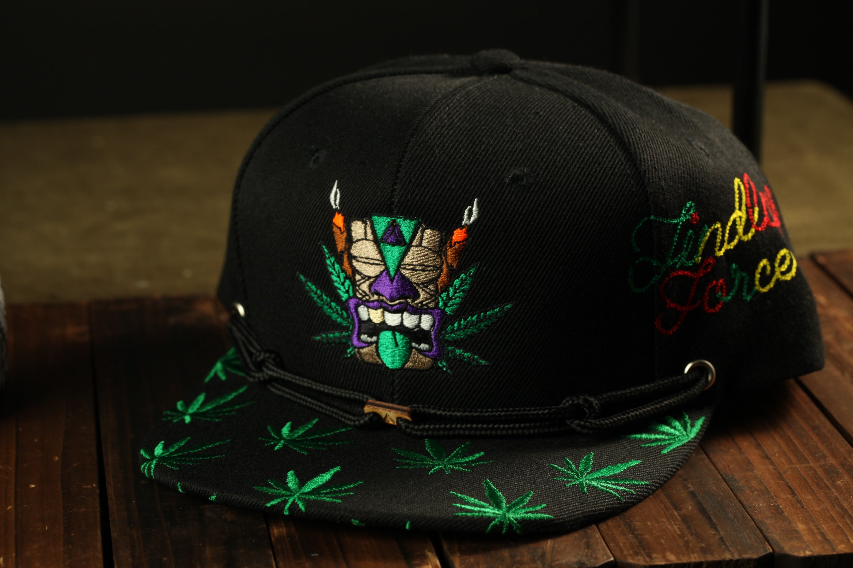 A black baseball cap with a colorful, embroidered design on the front, including a mask-like figure and green leaf patterns.