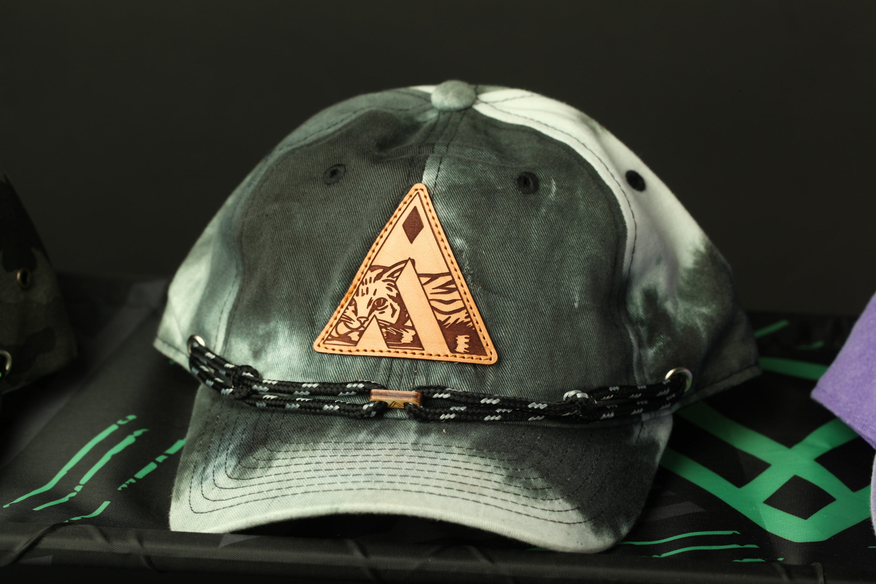 A gray and white baseball cap with a leather patch featuring a triangle design and a cat image, resting on a black surface.