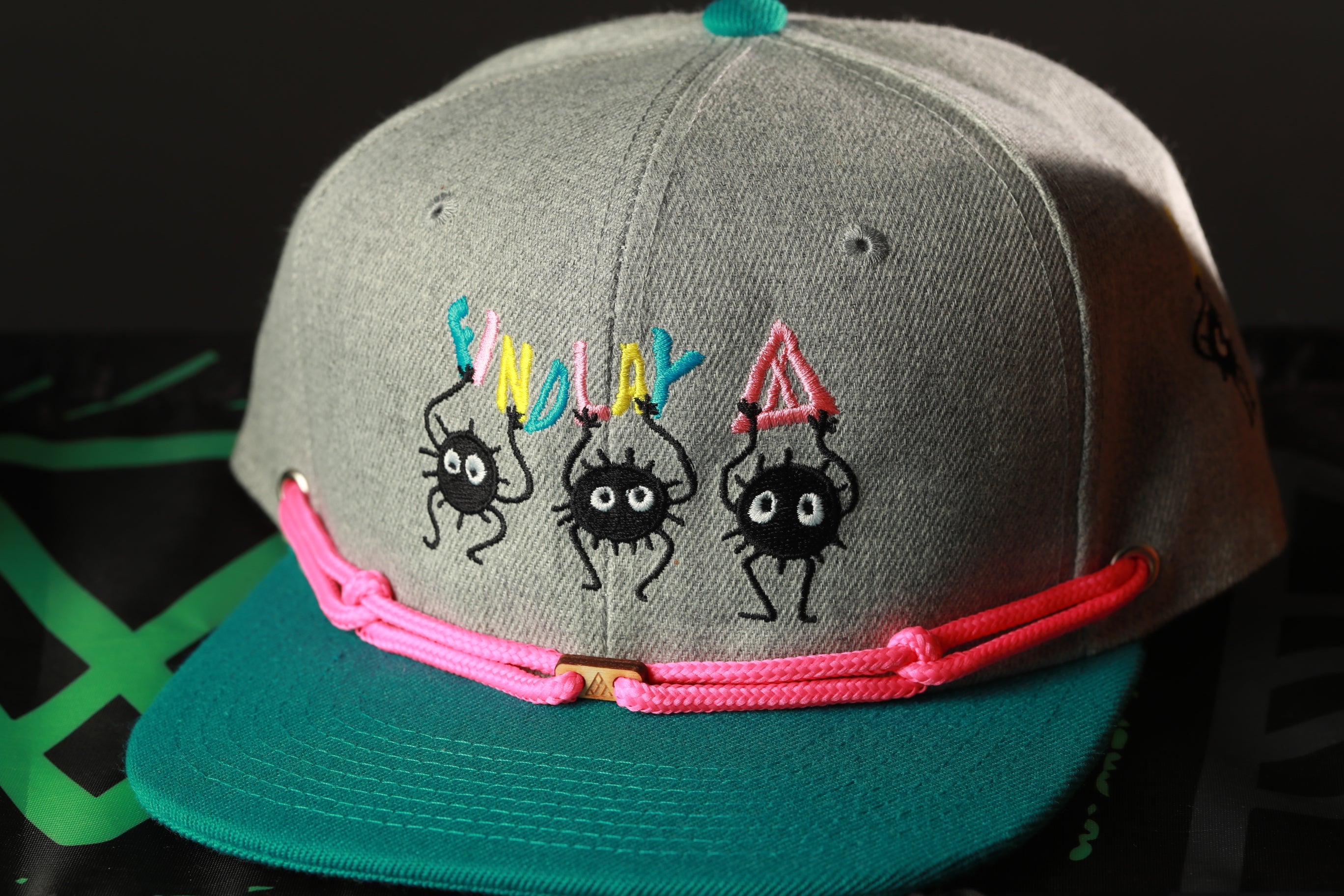 A gray baseball cap with colorful cartoon spider characters on it, and a pink string around the brim.