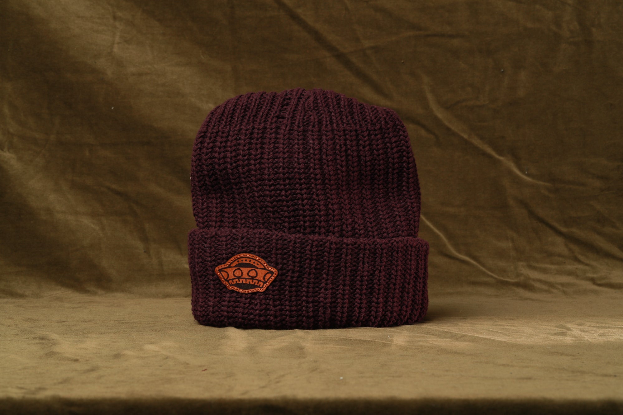 A burgundy knit beanie with an orange logo on the front, placed against a brown background.
