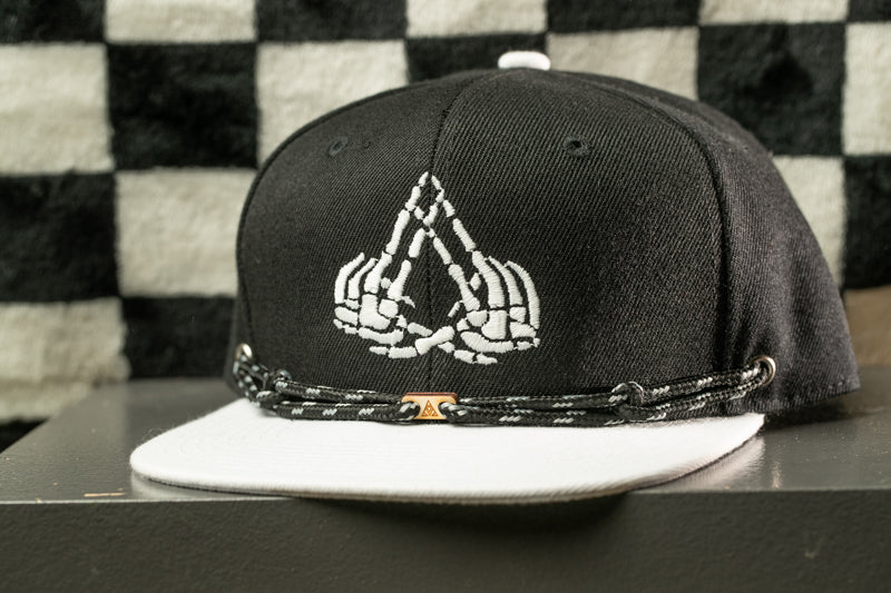 A black baseball cap with a white skull and crossbones design on the front, sitting on a white surface against a black and white checkered background.