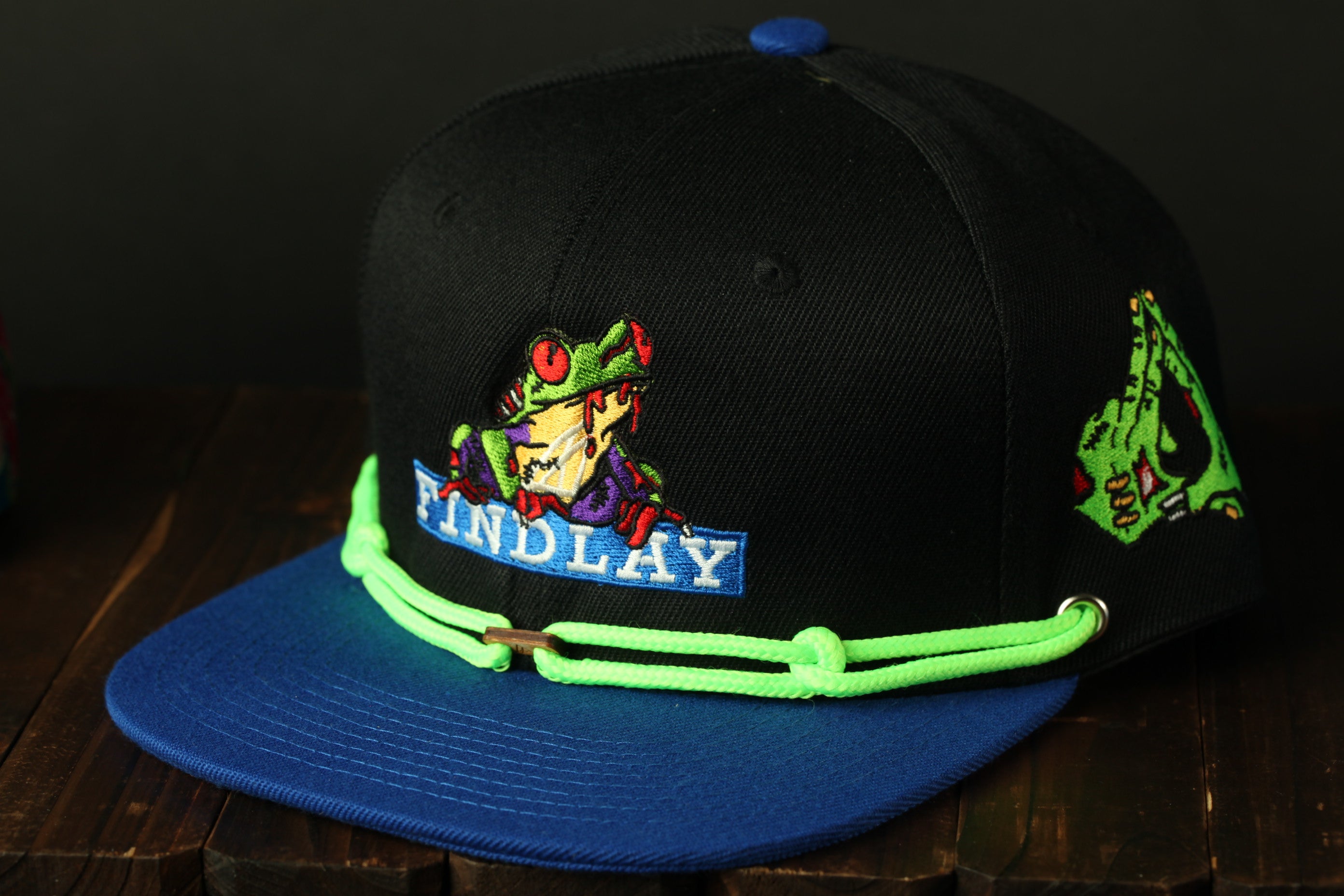 A black baseball cap with a frog design and the word "FINDLAY" printed on it, sitting on a wooden surface.