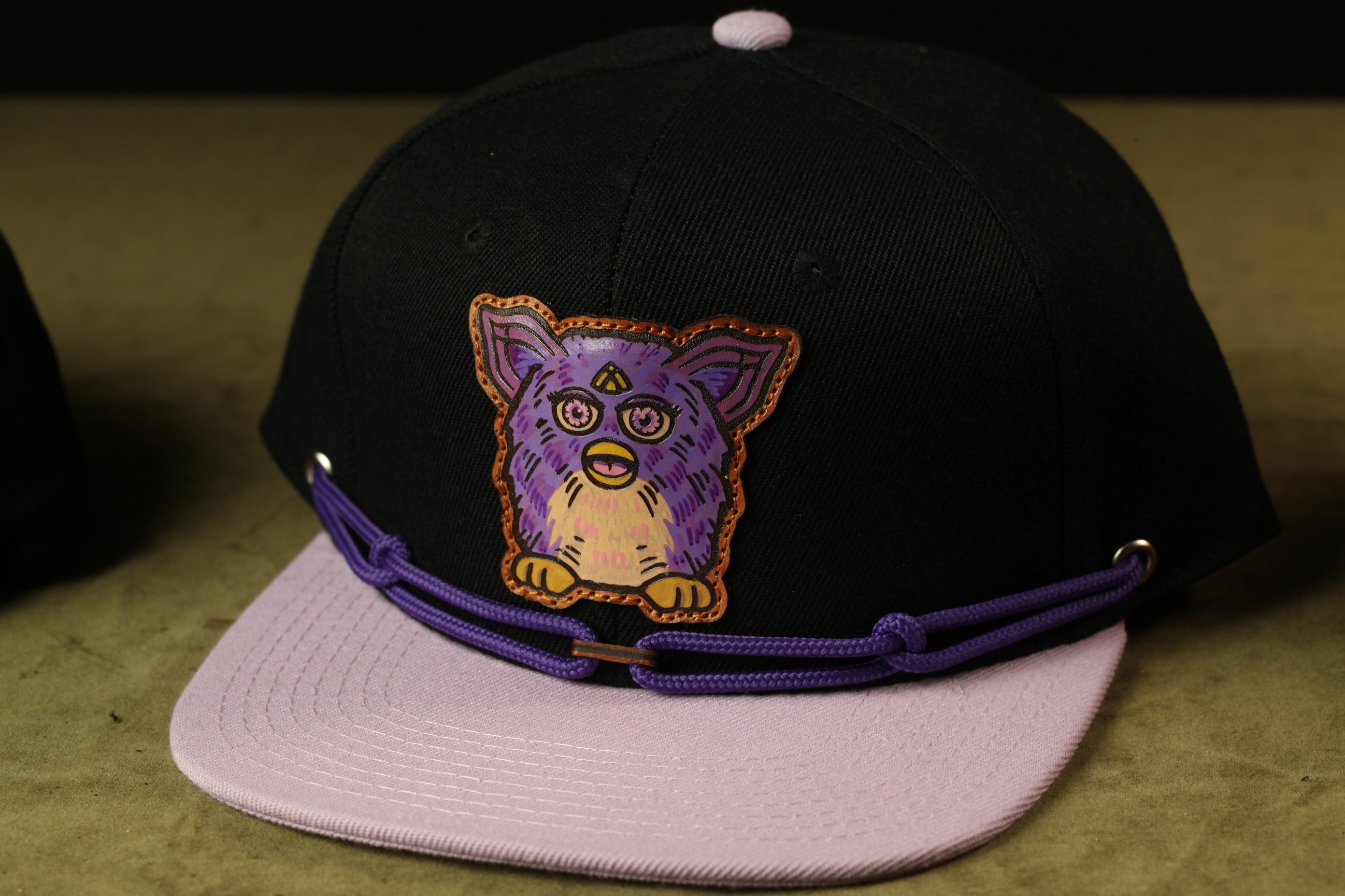A black baseball cap with a purple and orange design featuring a cartoon-like face on the front.