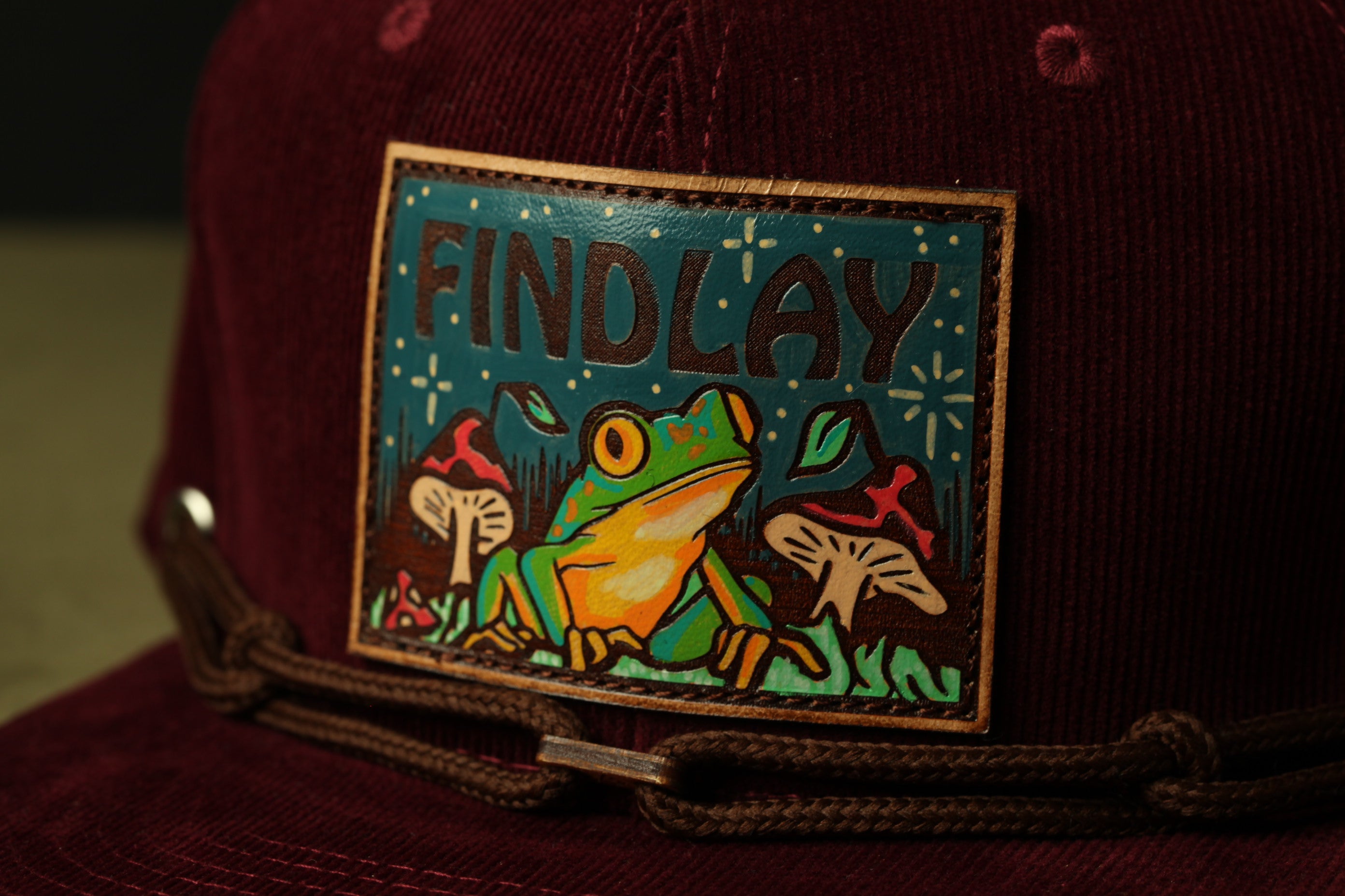 A close-up of a burgundy-colored baseball cap with a patch featuring a frog and mushrooms.