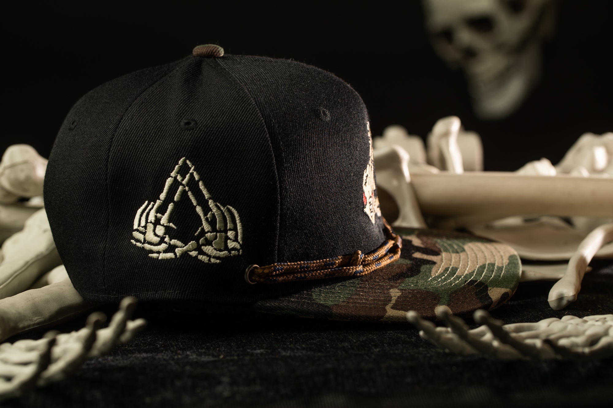 A black baseball cap with a white skull design is placed on a surface with various skeletal remains, including bones and a skeleton head.