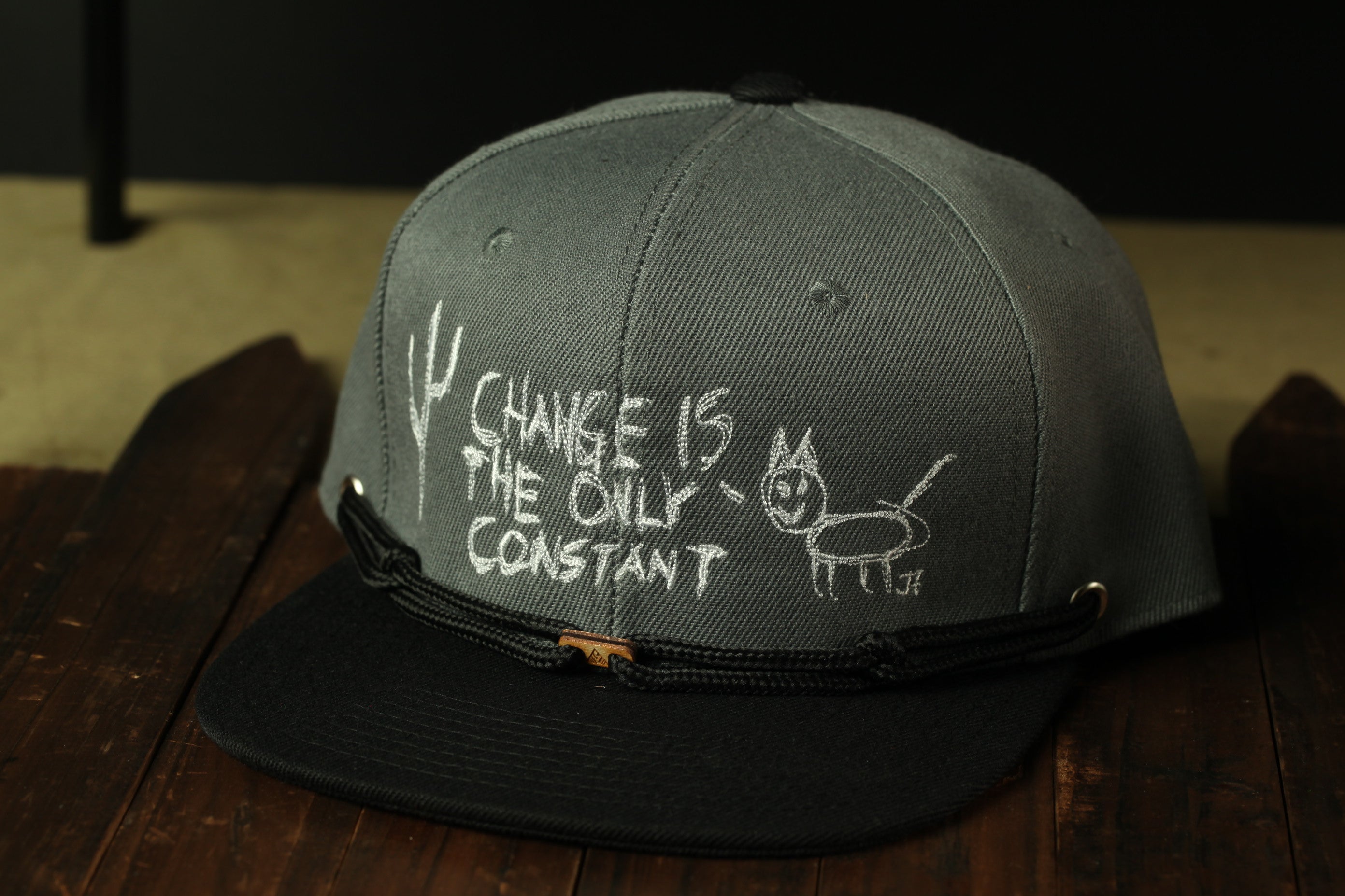 A gray baseball cap with white text and a drawing on it, placed on a wooden surface.