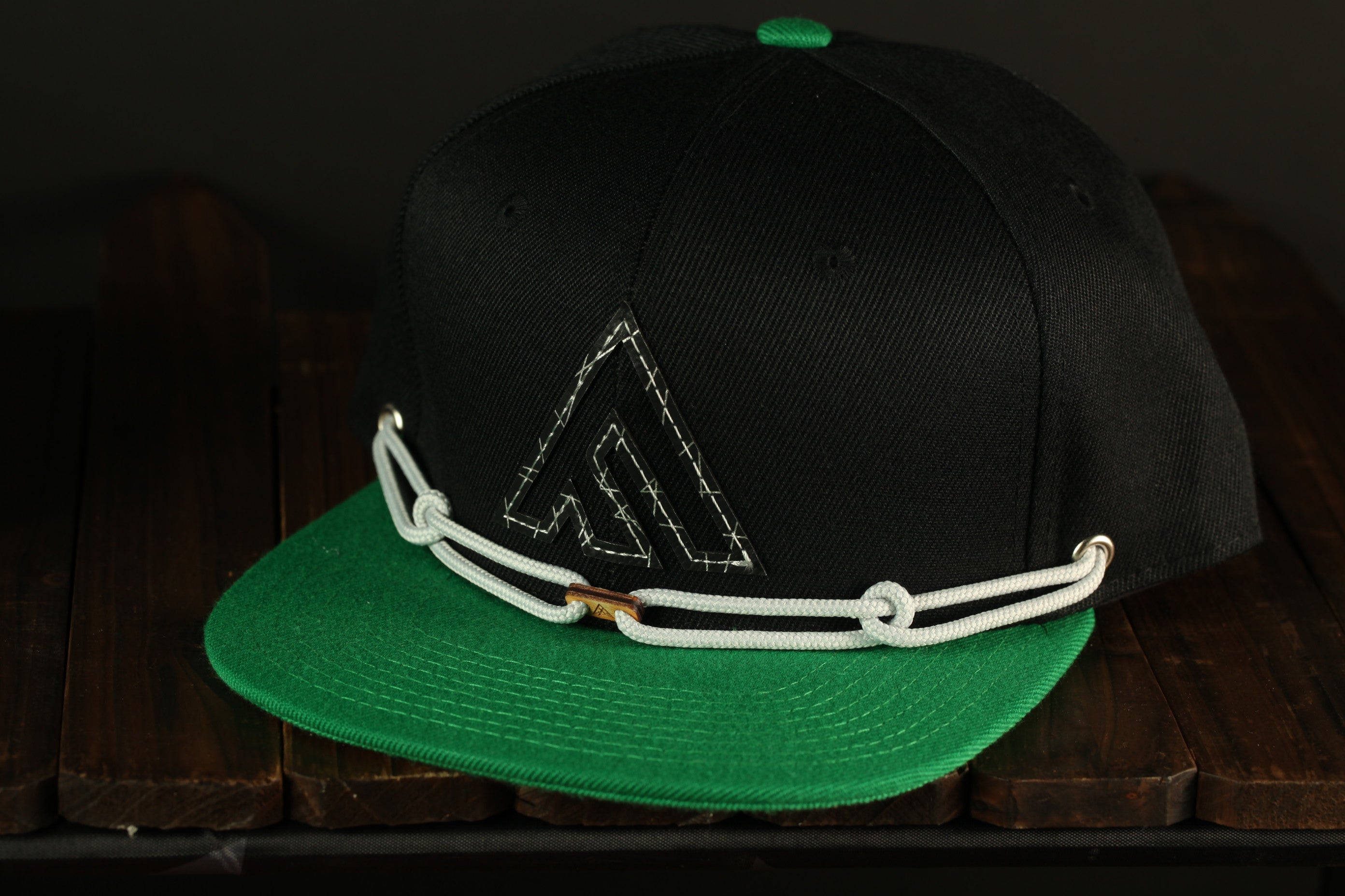 A black baseball cap with a green brim and a white rope strap is displayed on a wooden surface.