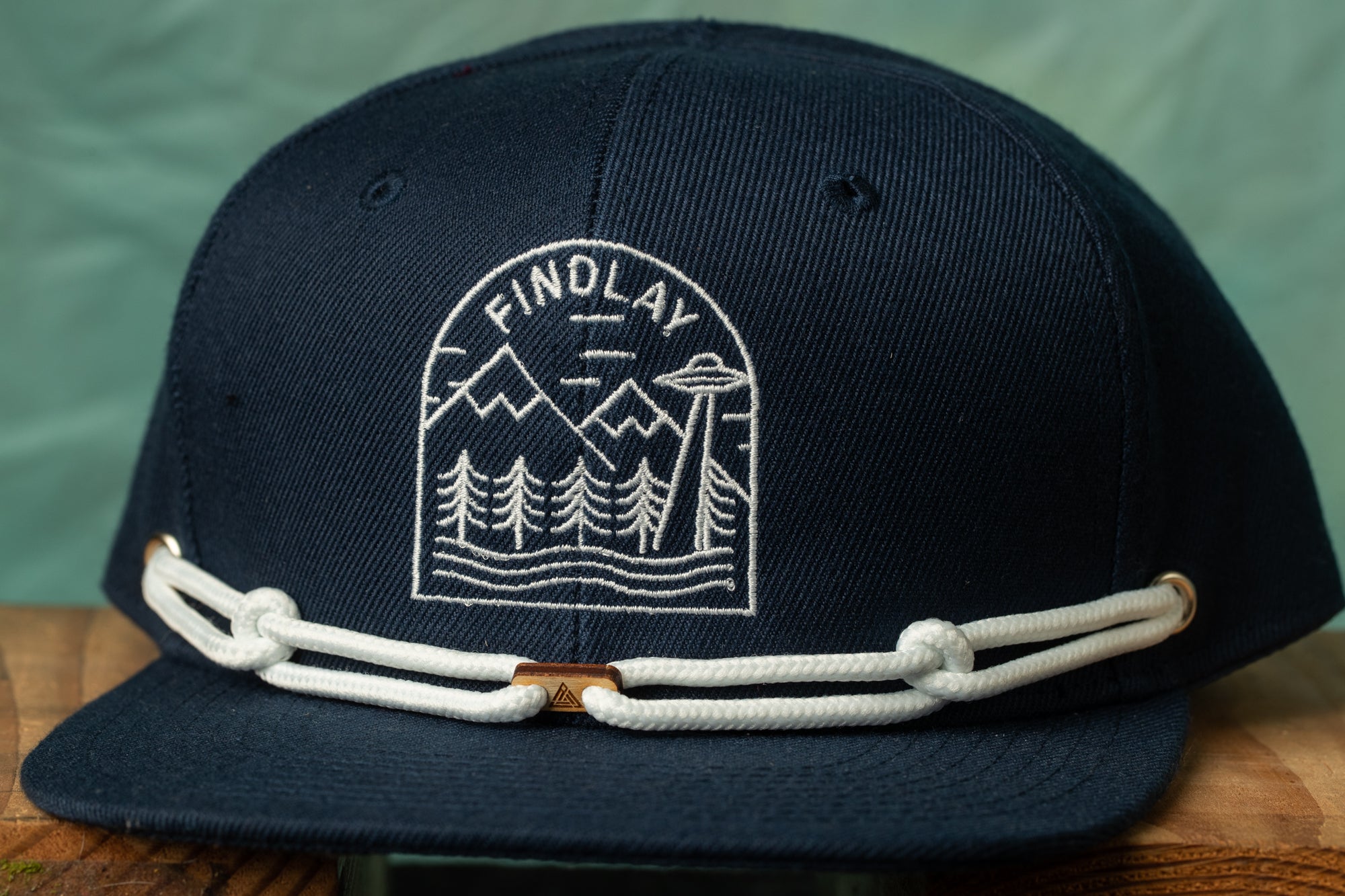 A navy blue baseball cap with a white embroidered design on the front, featuring a mountain landscape and an alien spacecraft.