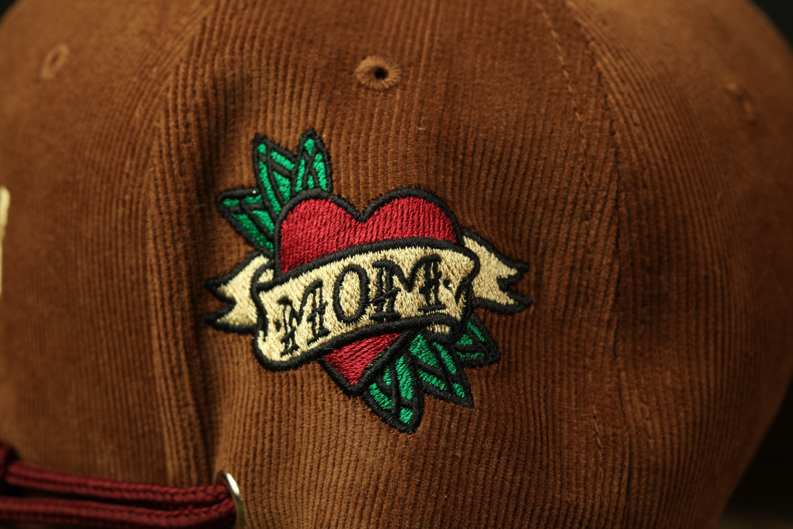 A close-up of a brown baseball cap with a heart-shaped embroidered patch that says "MOM" on it.