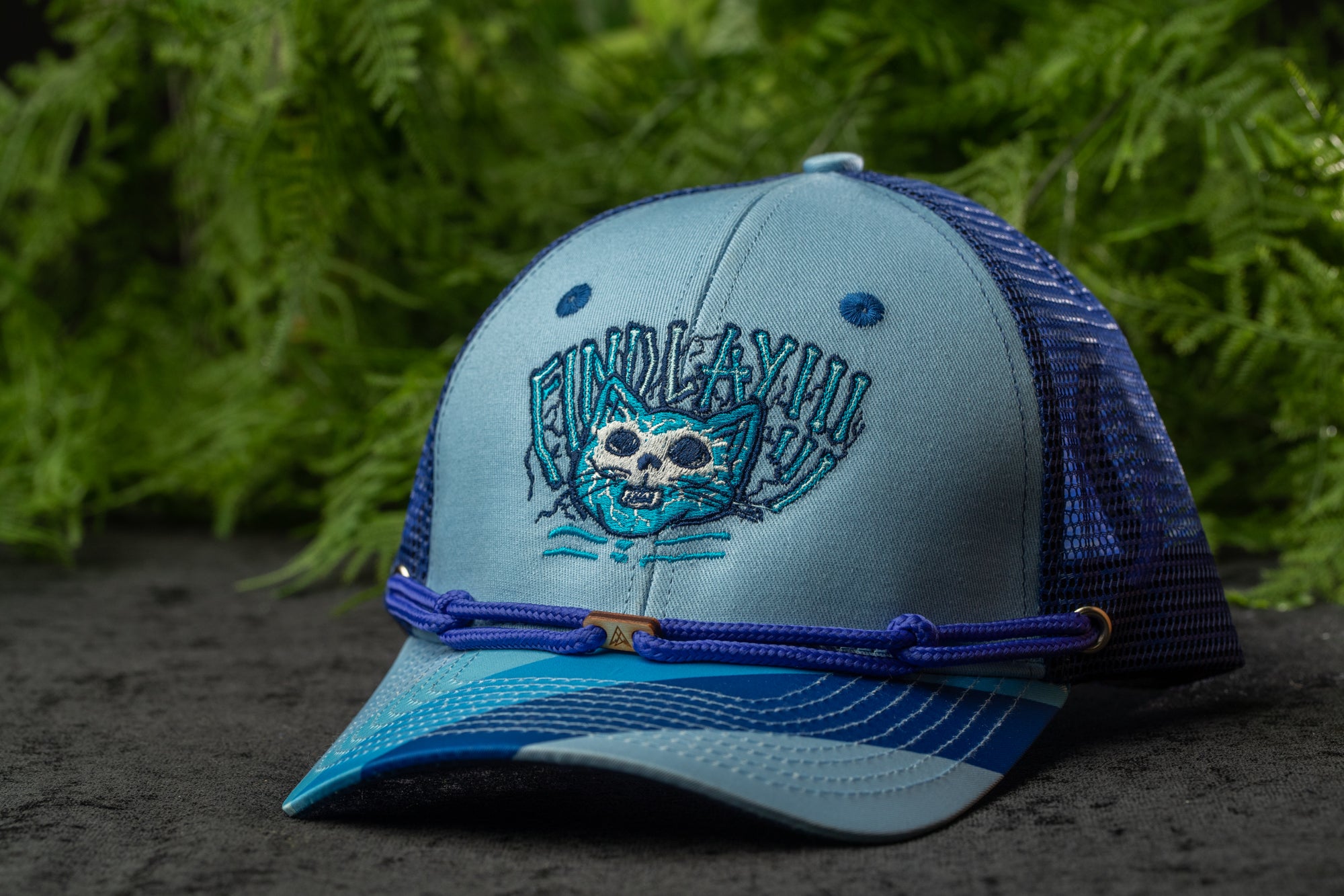 A blue and gray baseball cap with a cat design on it, placed on a surface with green foliage in the background.