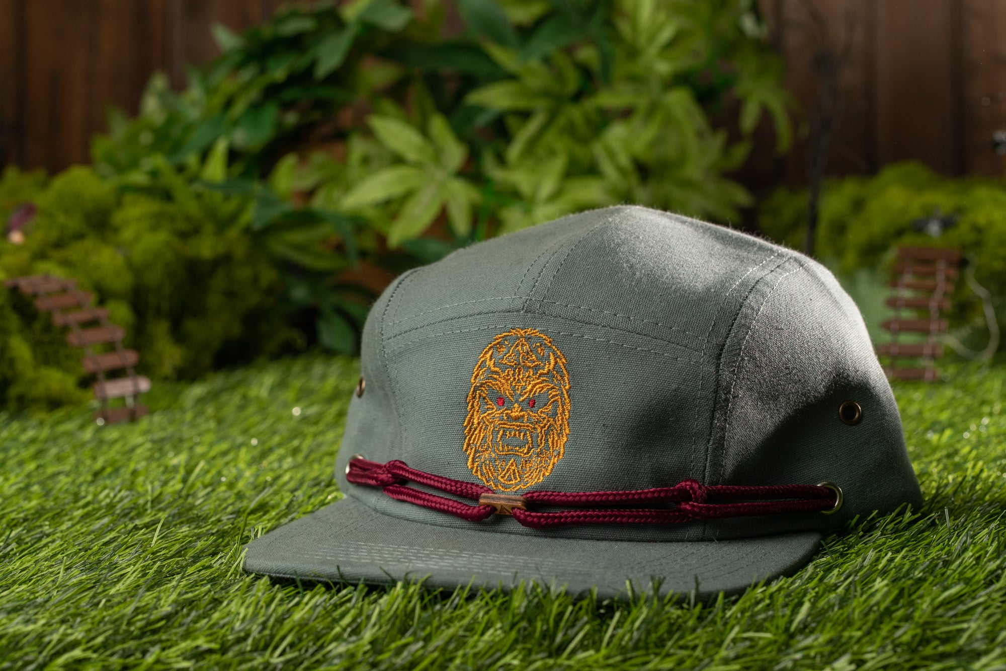 A green baseball cap with a red band and an embroidered design on the front, placed on a grassy surface with a blurred background of greenery.
