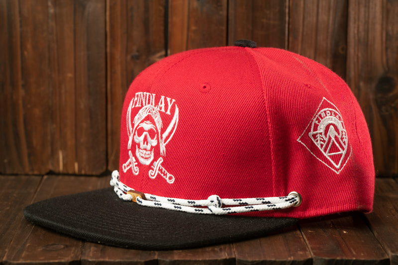A red baseball cap with a skull and crossbones design, featuring a black brim and a white rope trim, is displayed against a wooden background.