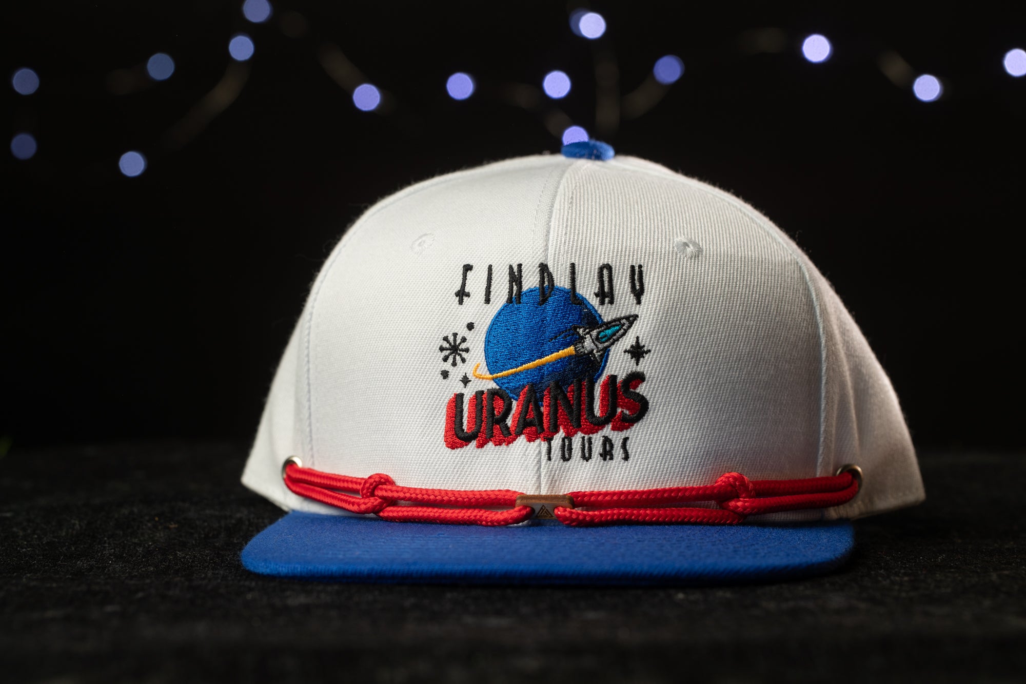 A white baseball cap with a blue brim and red strings, featuring a graphic of a planet and the text "Find Uranus Tours" on the front.