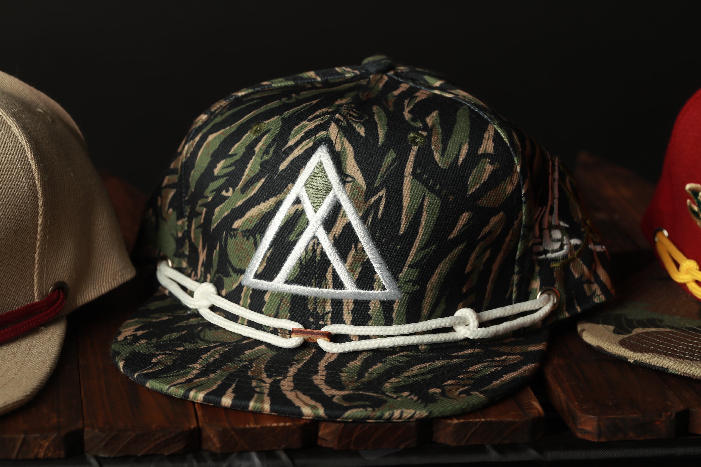 A camouflage baseball cap with a white triangle logo is displayed on a wooden surface, accompanied by other hats.