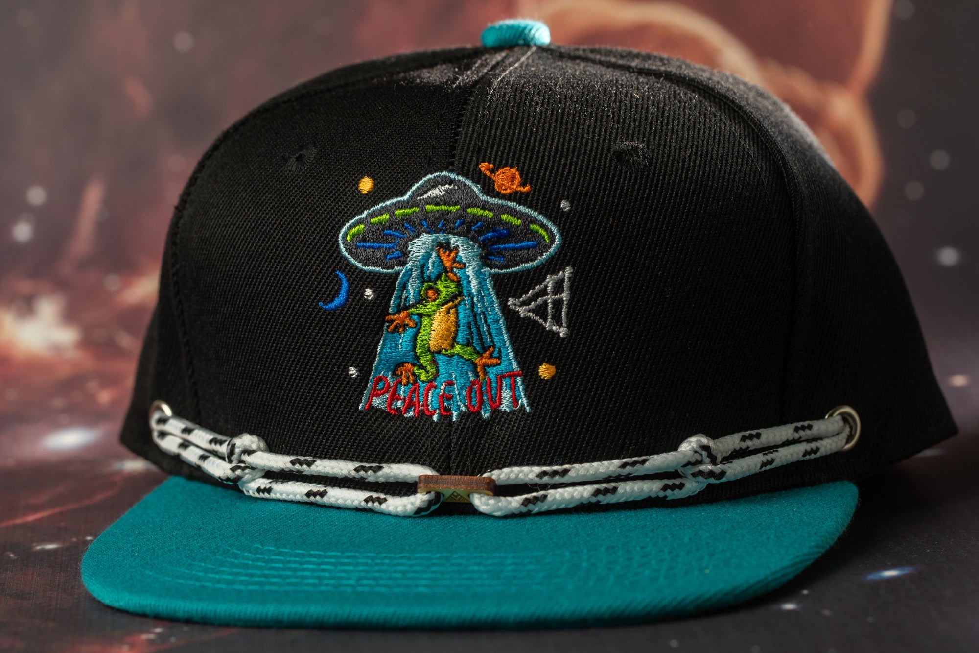 A black baseball cap with a colorful embroidered design on the front, featuring a green alien and the text "Peace Out" on a teal brim.