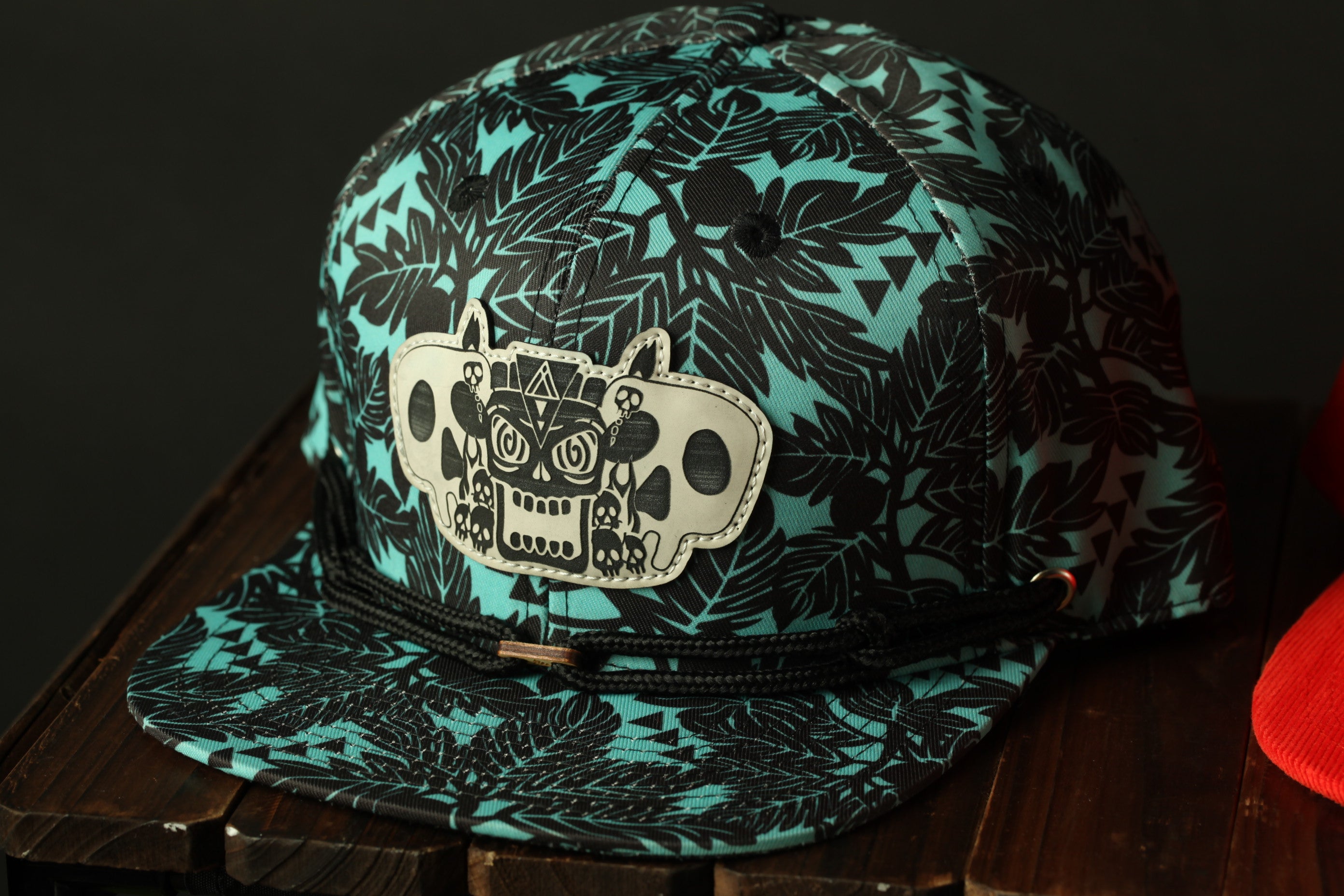 A turquoise baseball cap with a black leaf pattern and a skull design on the front, sitting on a wooden surface.