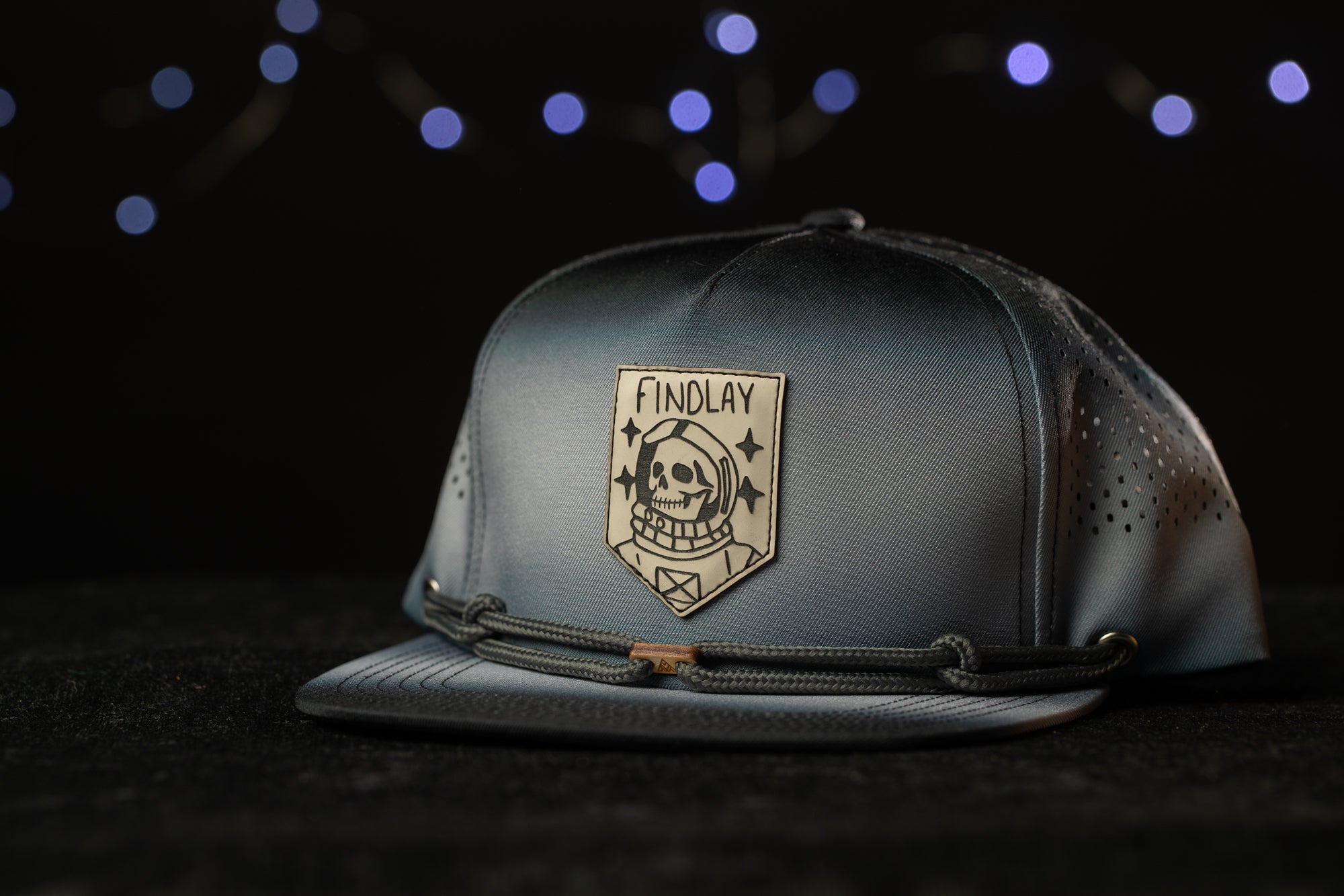 A gray baseball cap with a skull and crossbones design and the word "FINDLAY" printed on it, against a dark background with blue lights.