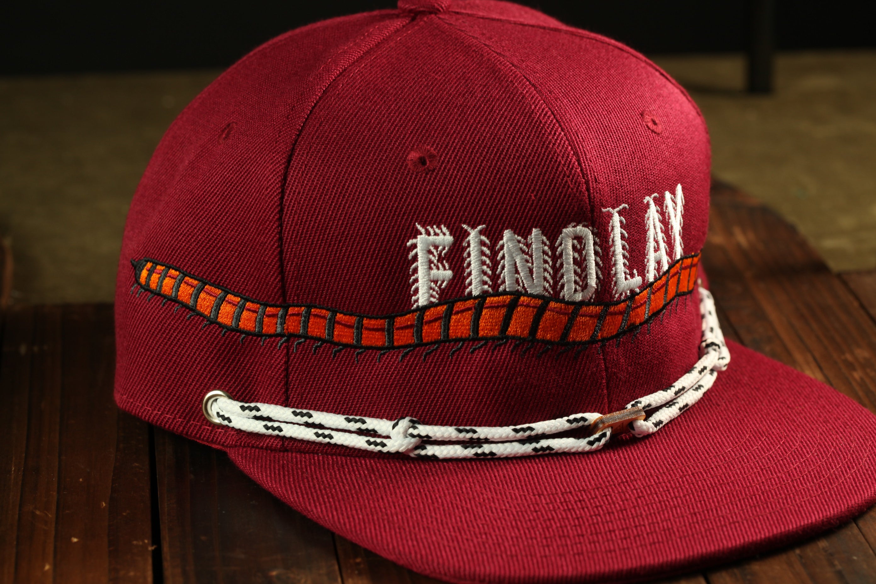 A maroon baseball cap with the word "FINOLI" embroidered on it, sitting on a wooden surface.