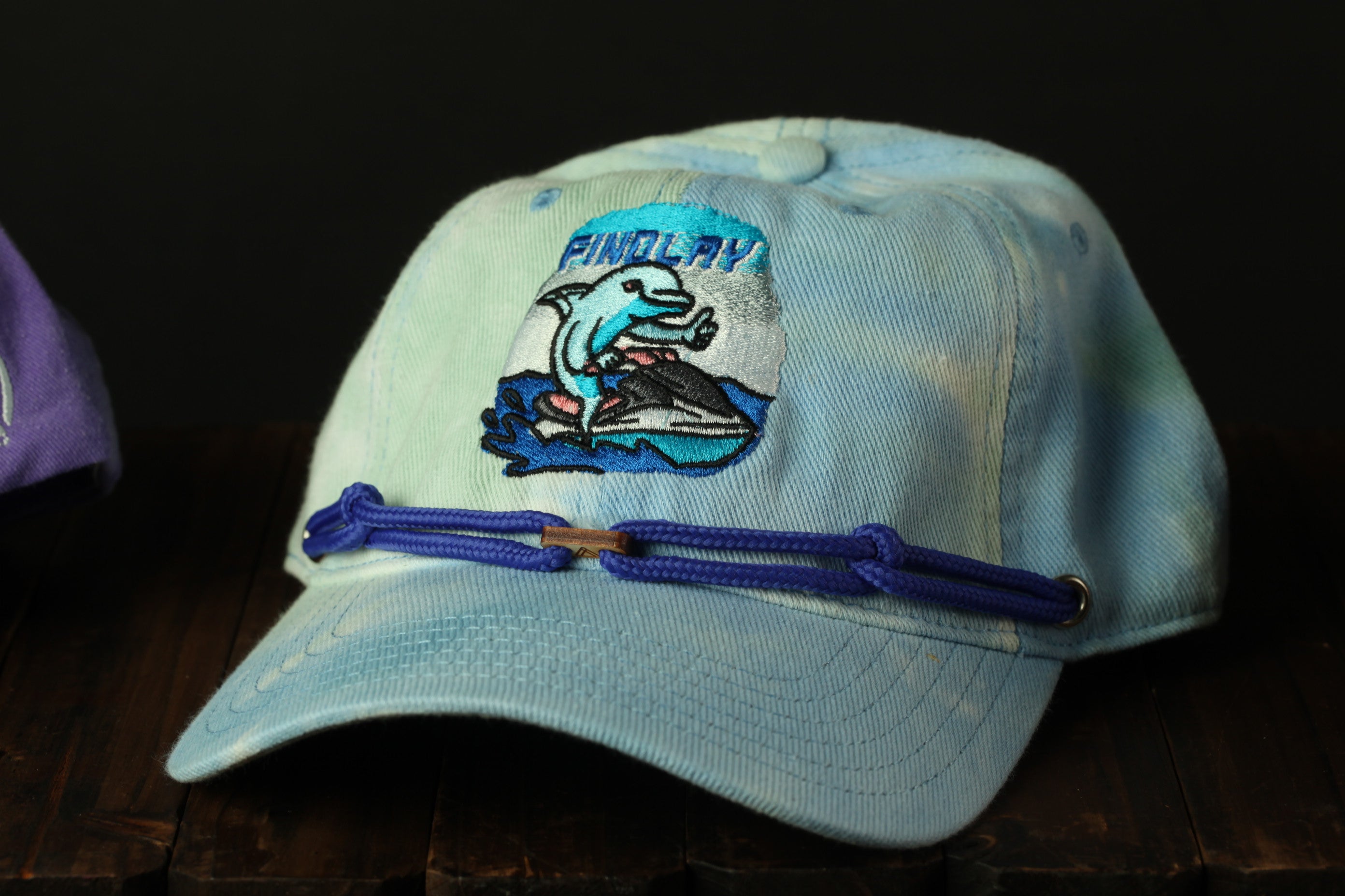 A blue baseball cap with a tie-dye design and a cartoon dolphin on it, sitting on a wooden surface.