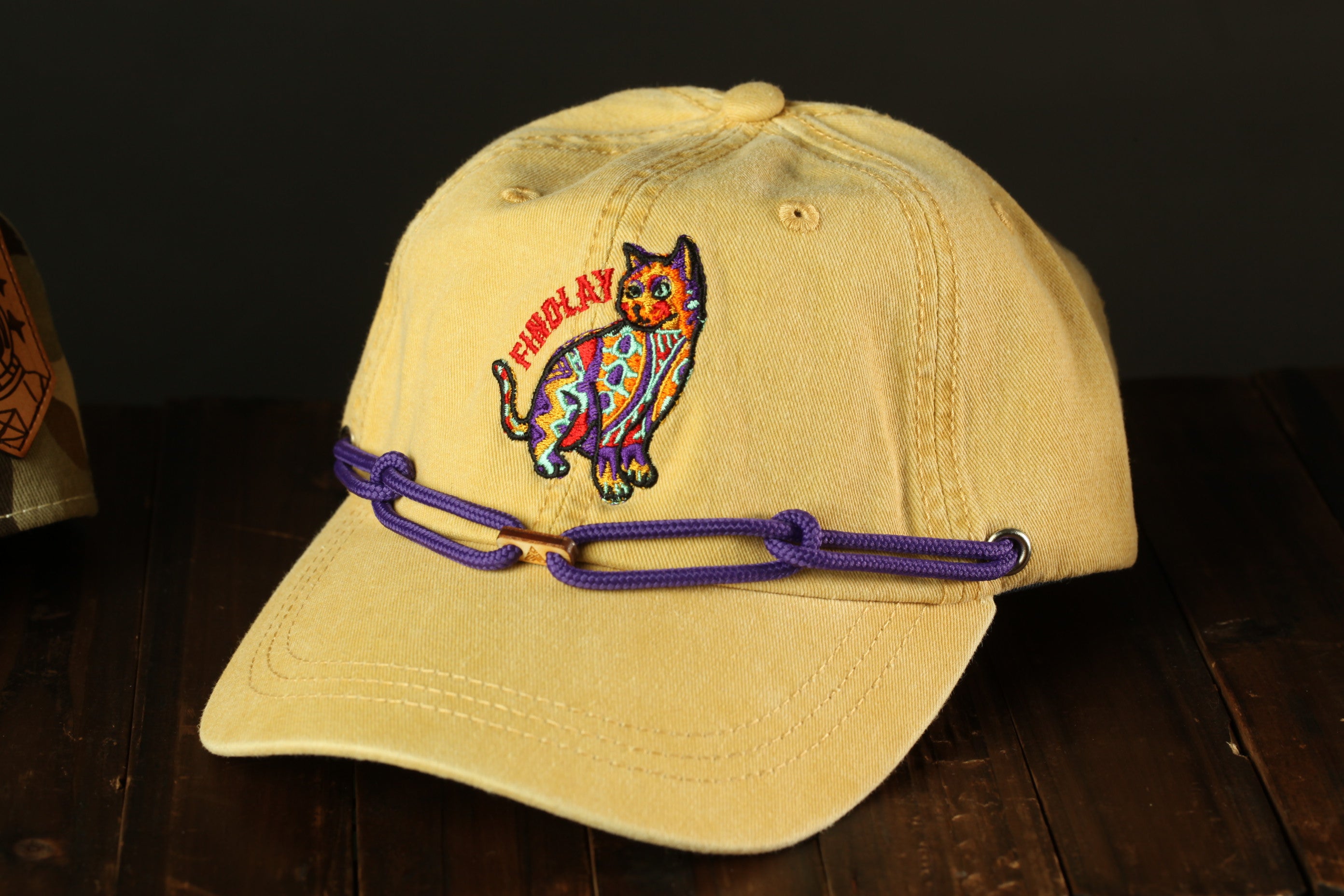 A yellow baseball cap with a colorful cat embroidered on it, sitting on a wooden surface.