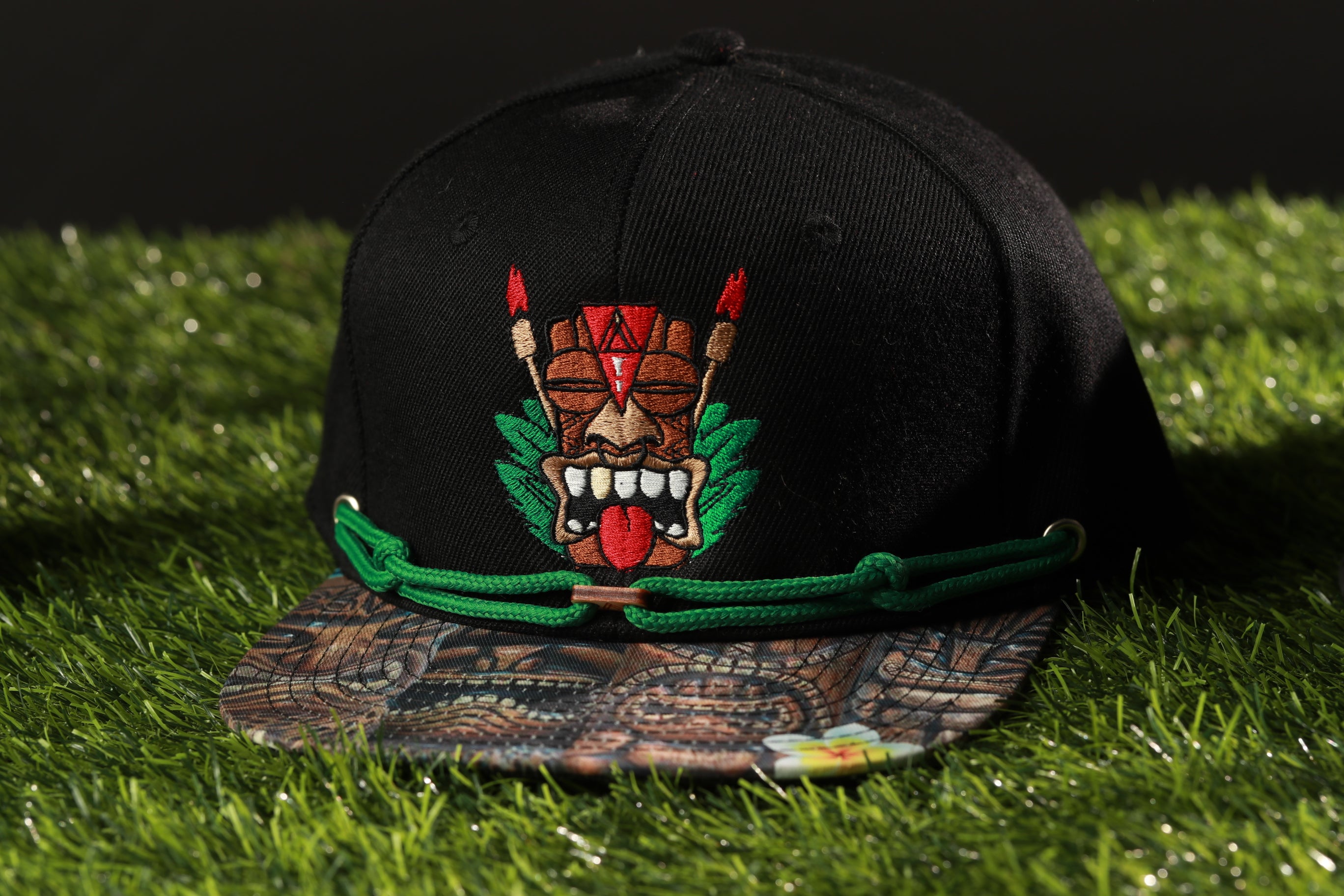 A black baseball cap with a colorful tiki mask design on the front, placed on a grassy surface.
