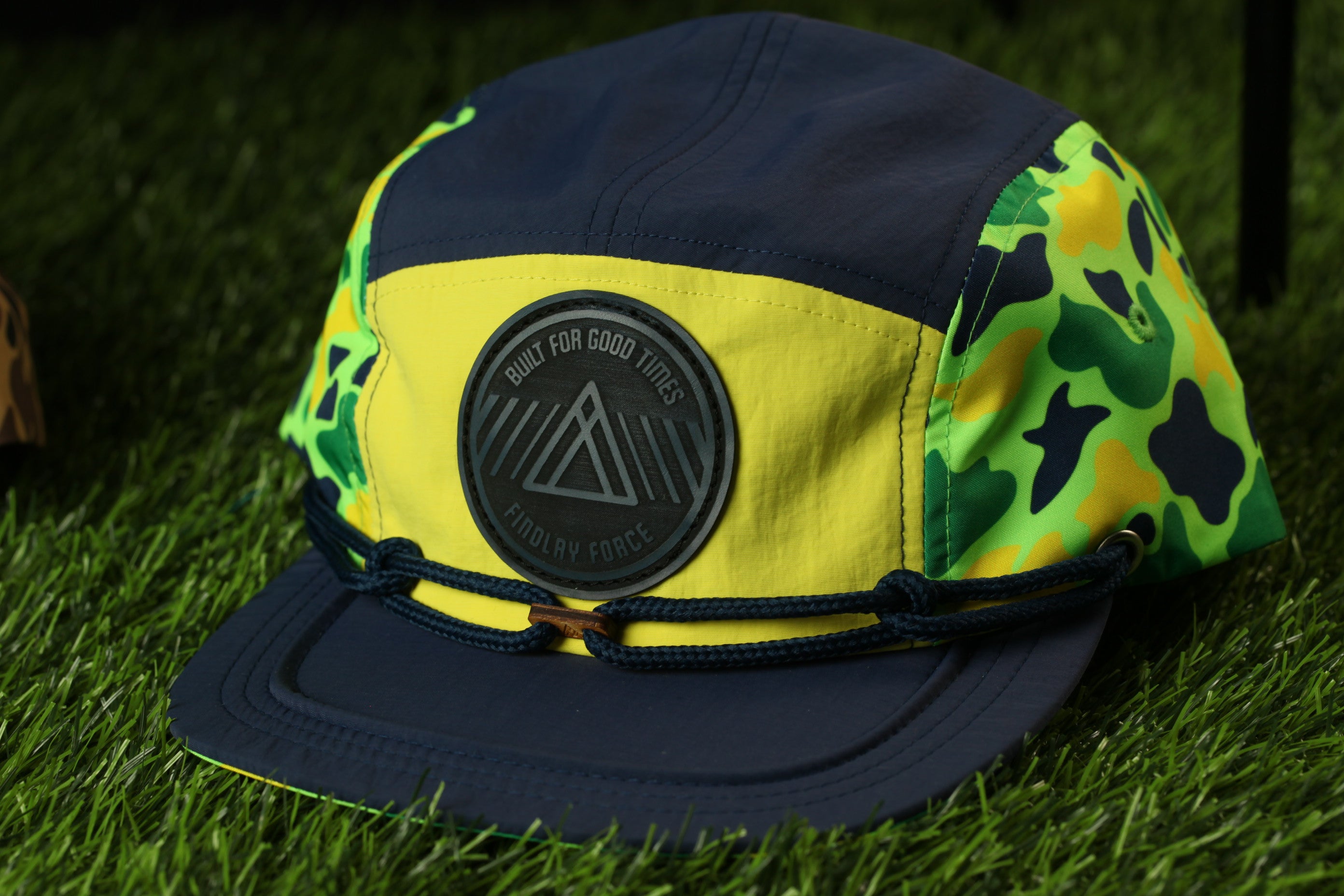 A yellow and blue baseball cap with a camouflage pattern and a black circular patch on the front, sitting on a grassy surface.