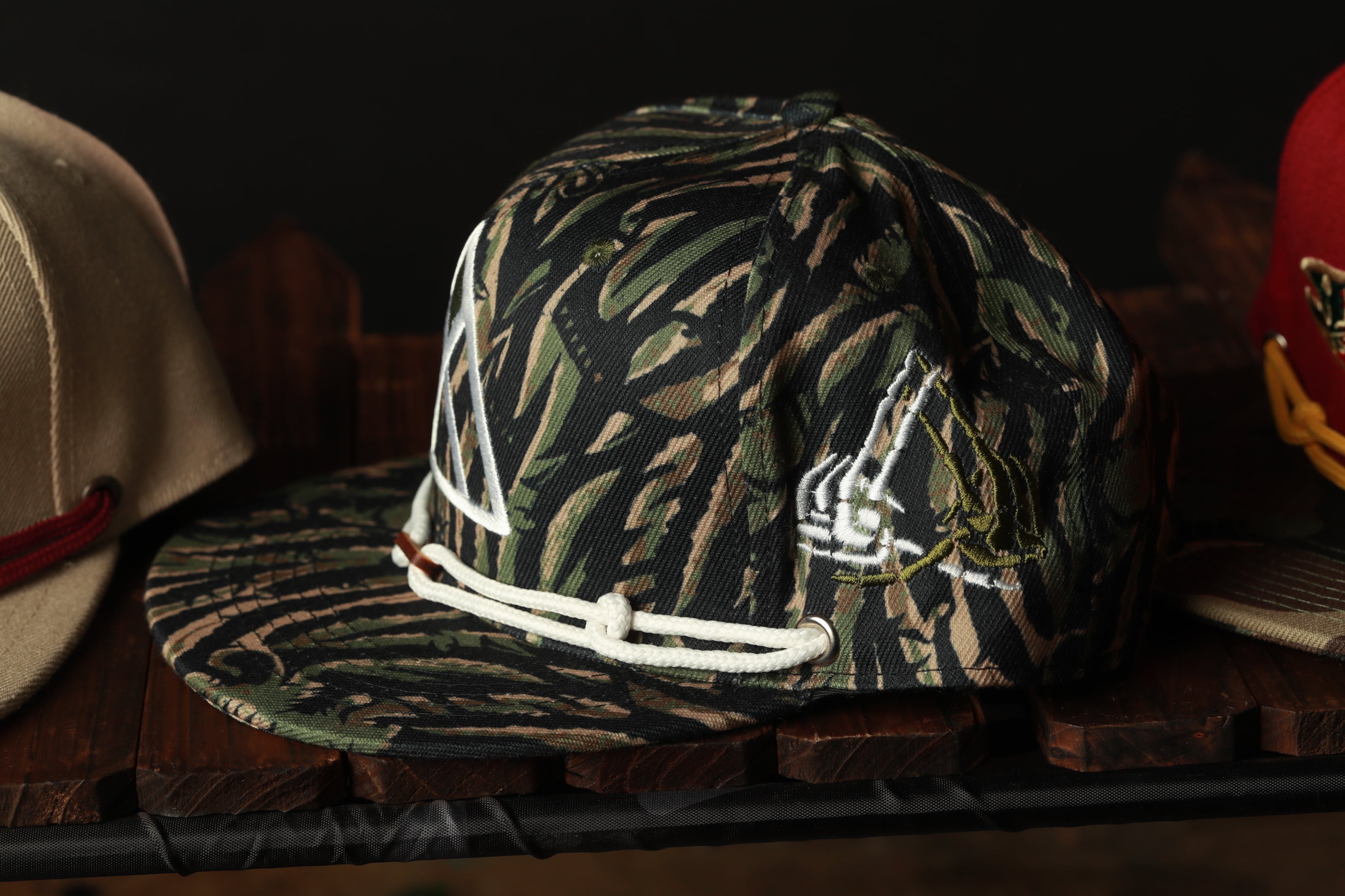 A camouflage baseball cap with a white logo is displayed on a wooden shelf, with other hats visible in the background.
