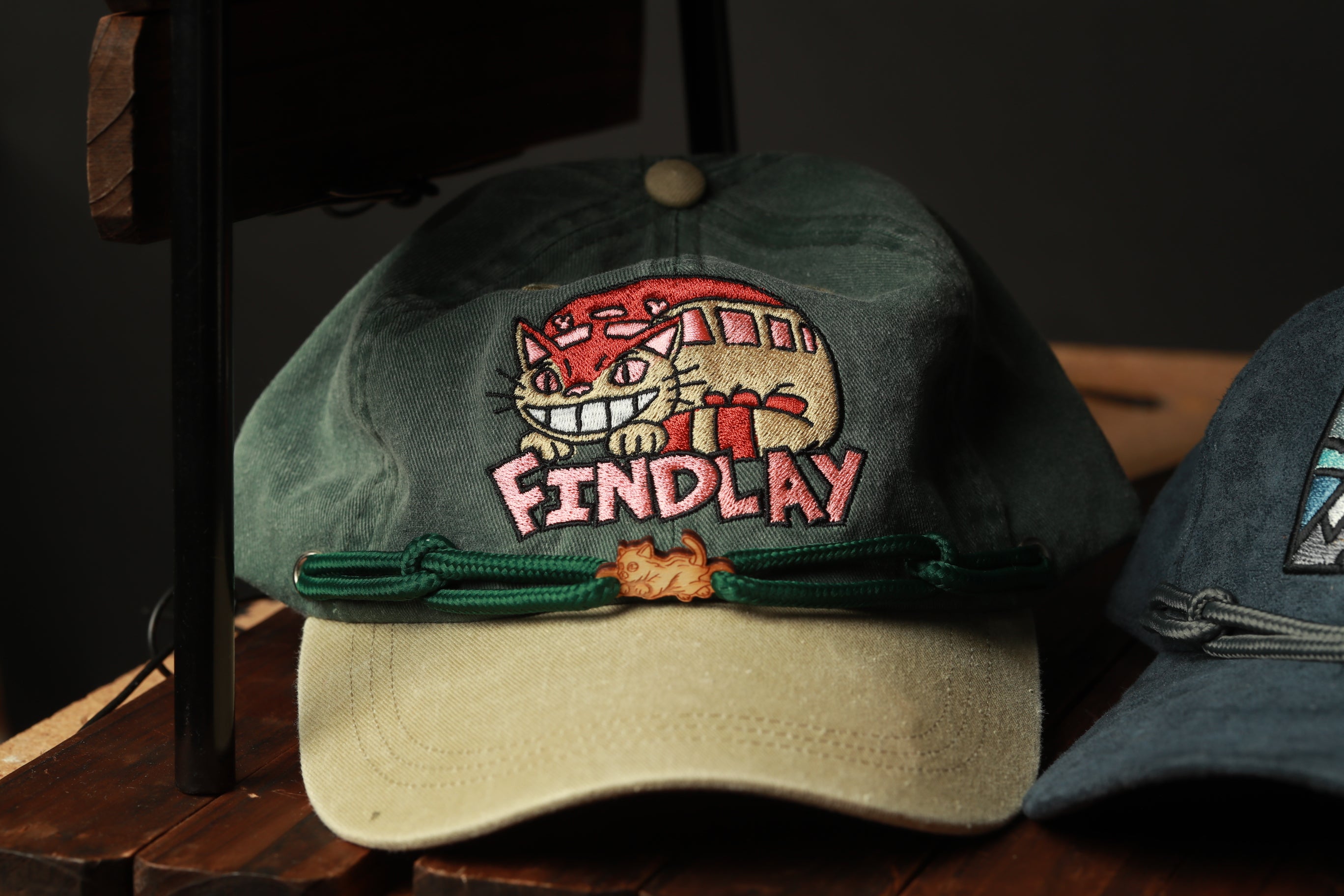 A green baseball cap with a cartoon cat design and the word "FINDlay" printed on it.