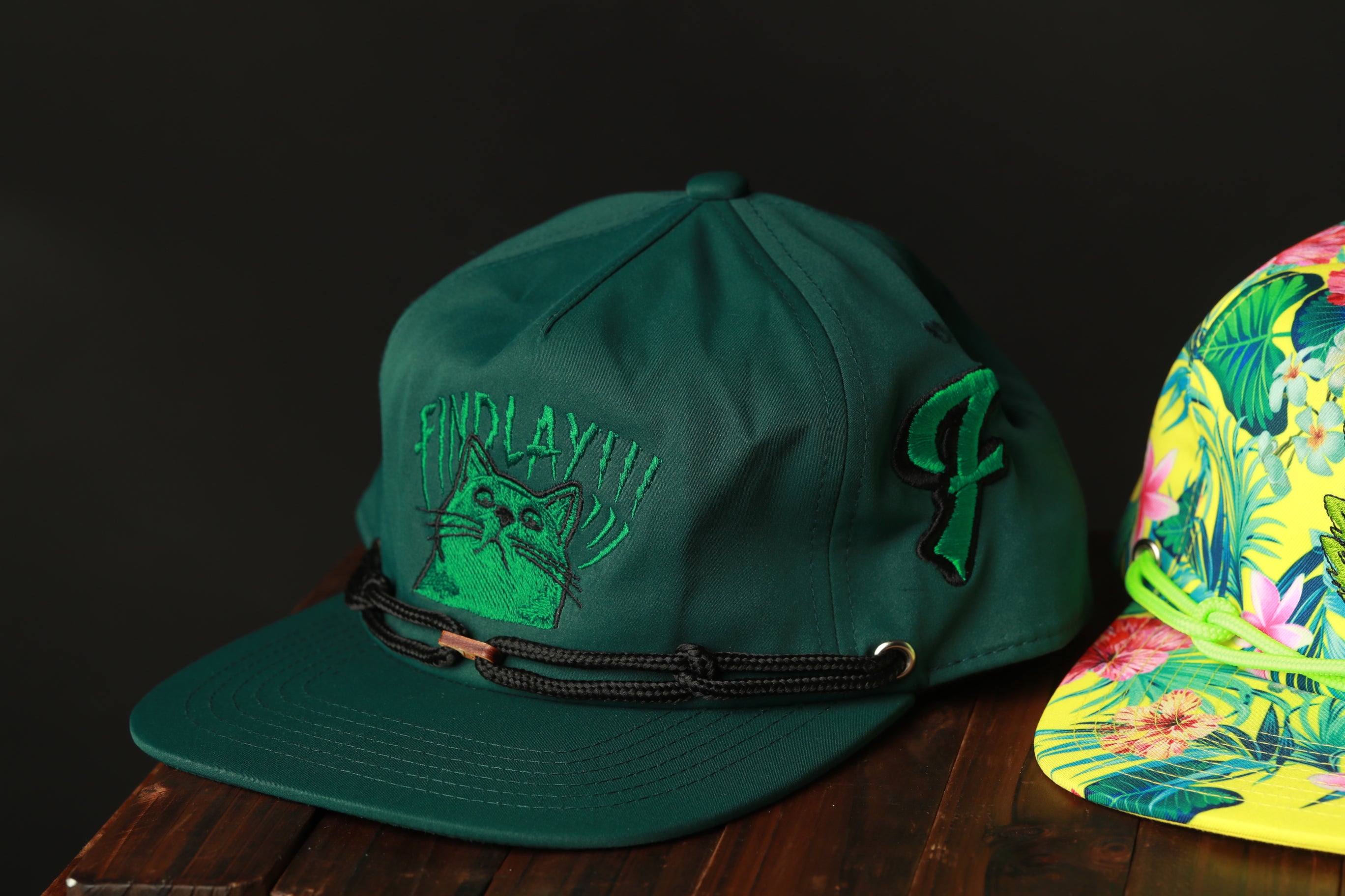 A green baseball cap with a cat embroidered on it, placed on a wooden surface next to another colorful cap.