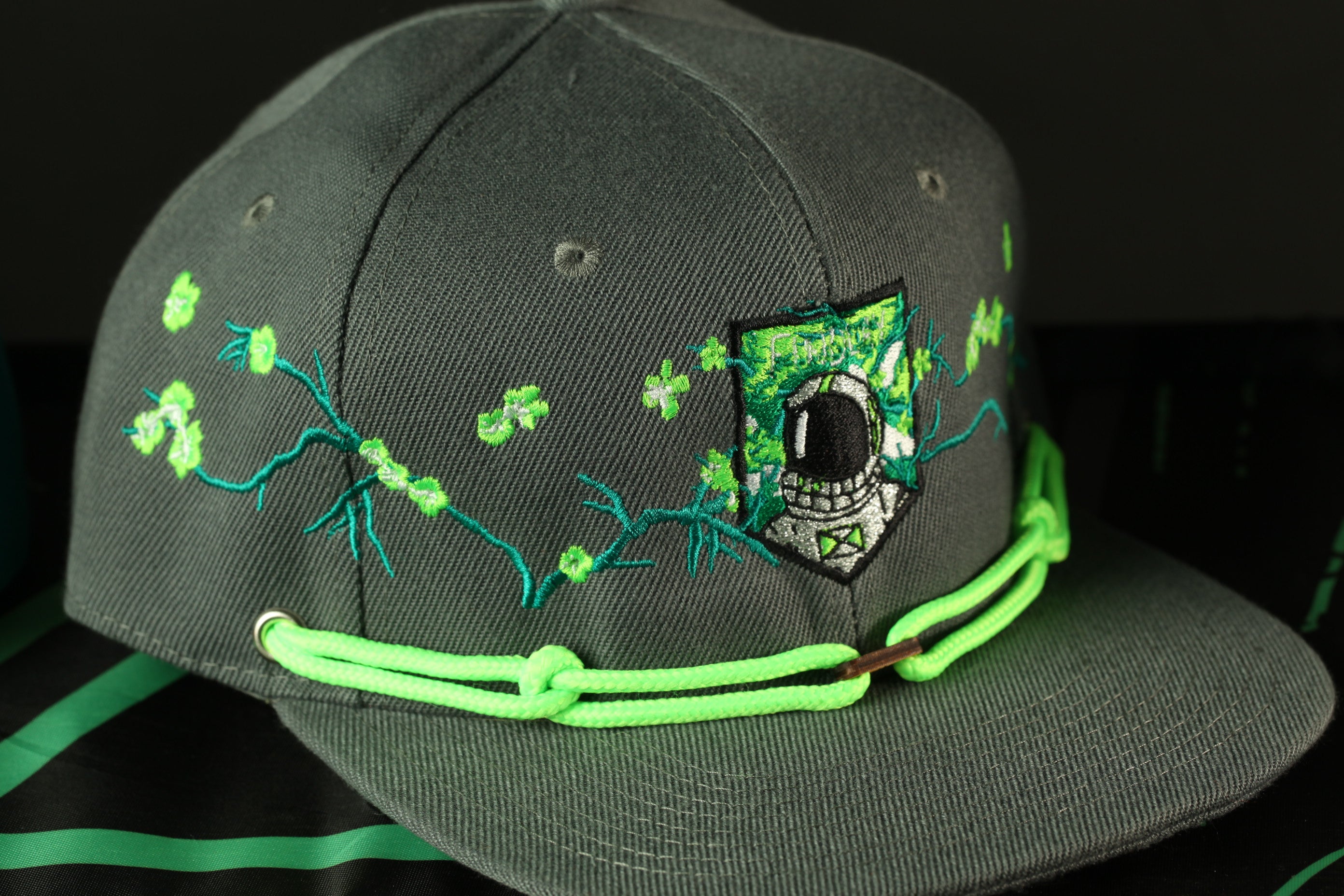 A gray baseball cap with green strings and a patch featuring a space suit and green leaves.