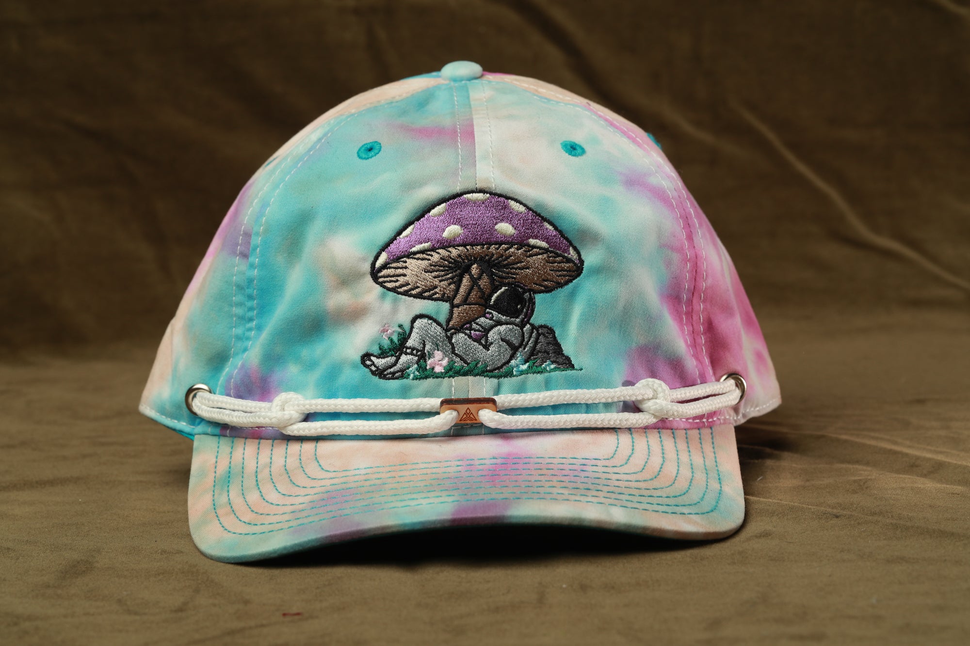 A tie-dye baseball cap with a mushroom design and a person sitting on a mushroom.