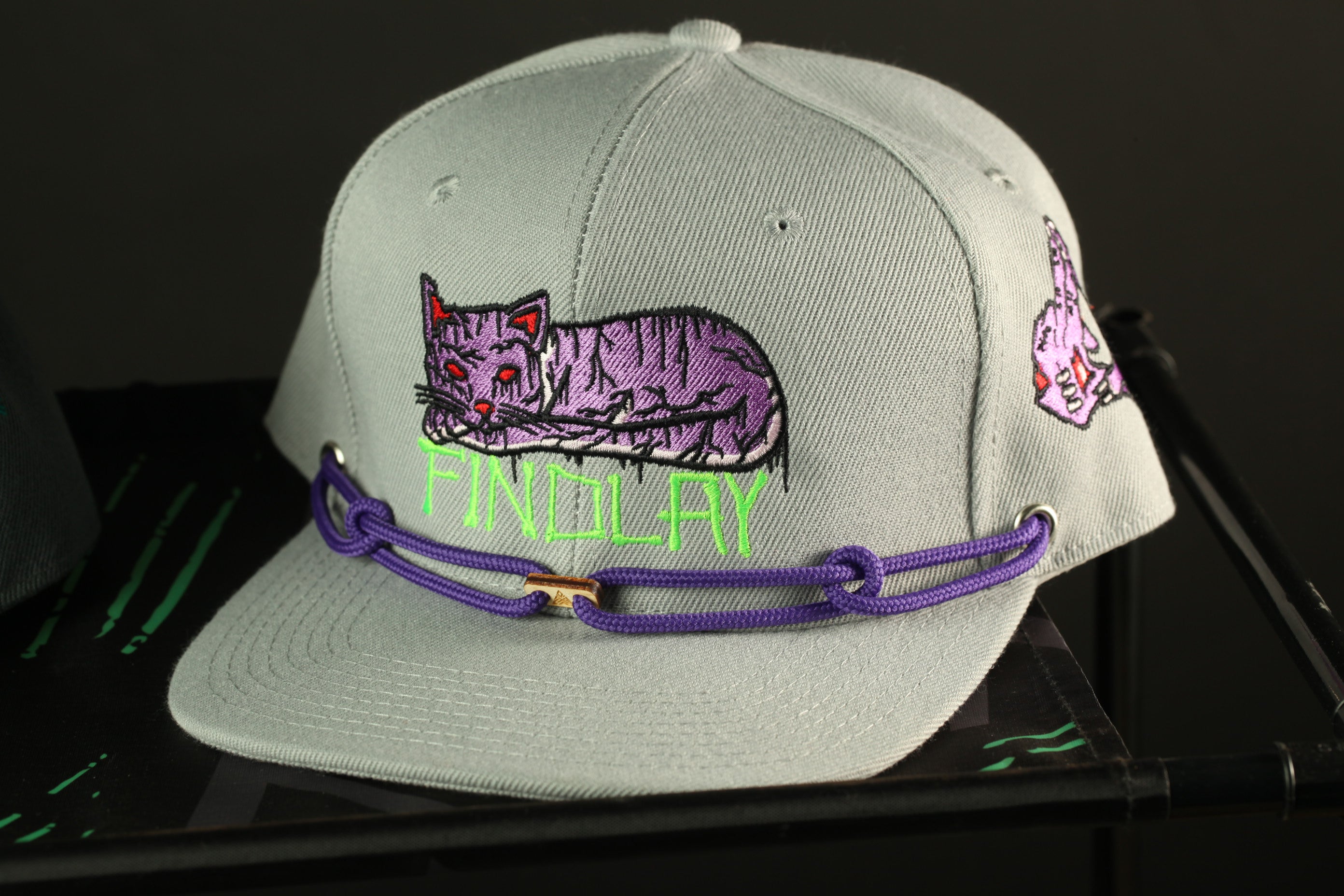 A gray baseball cap with a purple cat embroidered on it, and a purple string or strap attached to the brim.