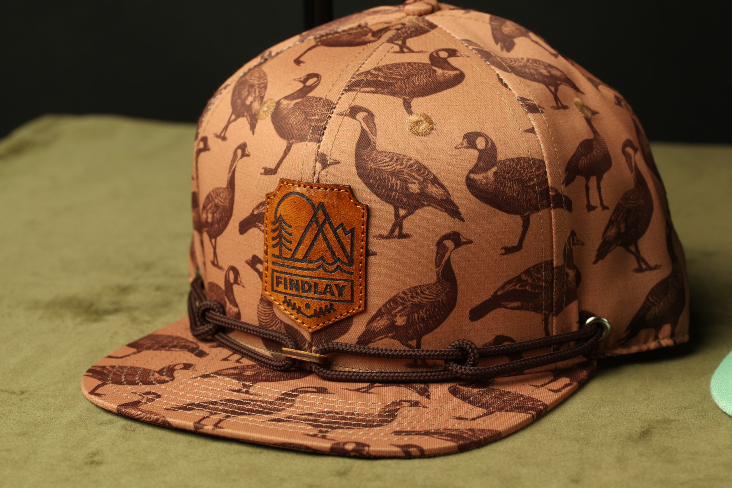 A brown baseball cap with a pattern of black and white birds printed on it, featuring a leather patch with the text "FINDLAY" and a mountain silhouette.
