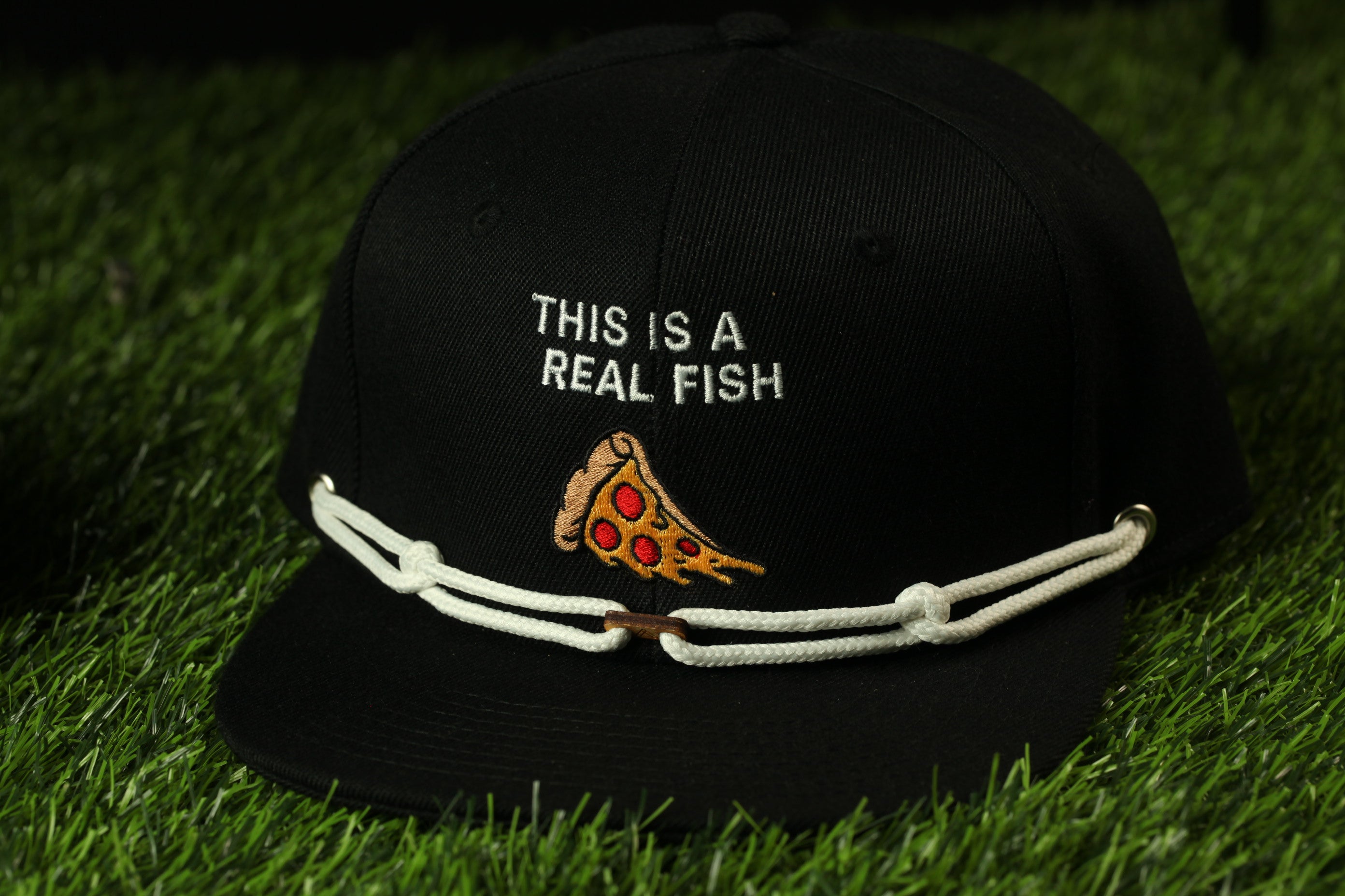 A black baseball cap with a white string and a pizza slice embroidered on it, placed on a grassy surface.