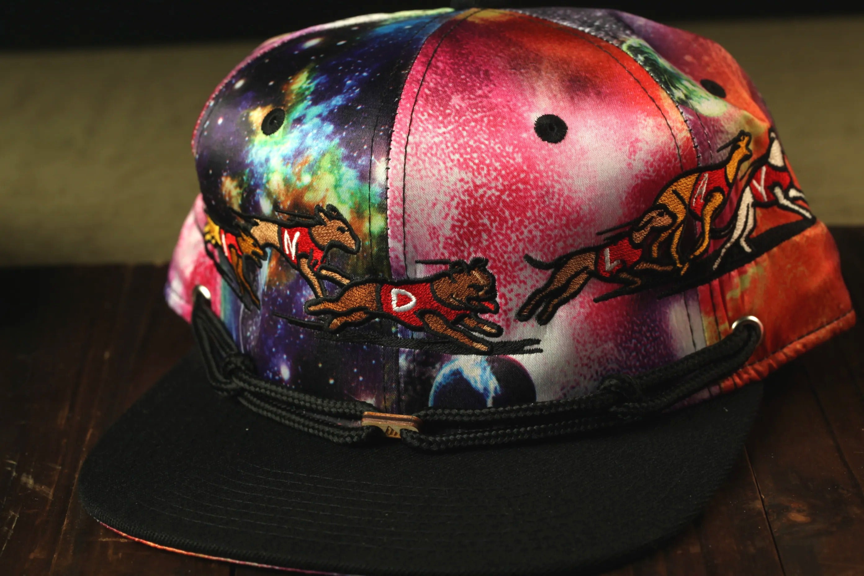 A colorful, patterned baseball cap with a space-themed design, featuring embroidered images of dogs running across a cosmic background.
