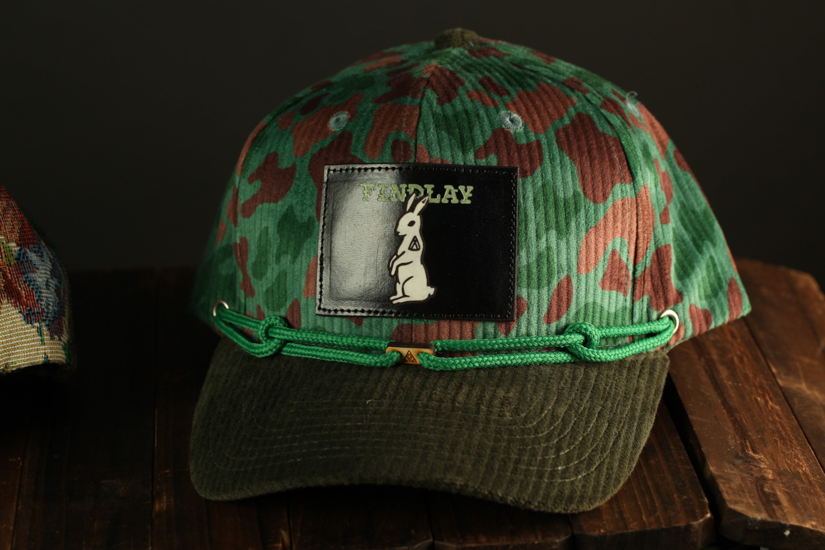 A camouflage-patterned baseball cap with a patch featuring a cartoon rabbit and the word "FIND LAY" on a wooden surface.