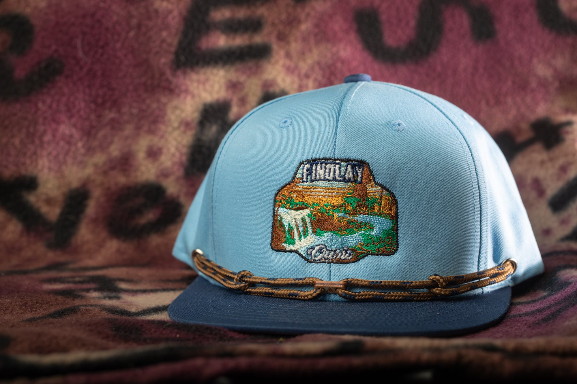A light blue baseball cap with a decorative patch on the front, featuring a scenic landscape.