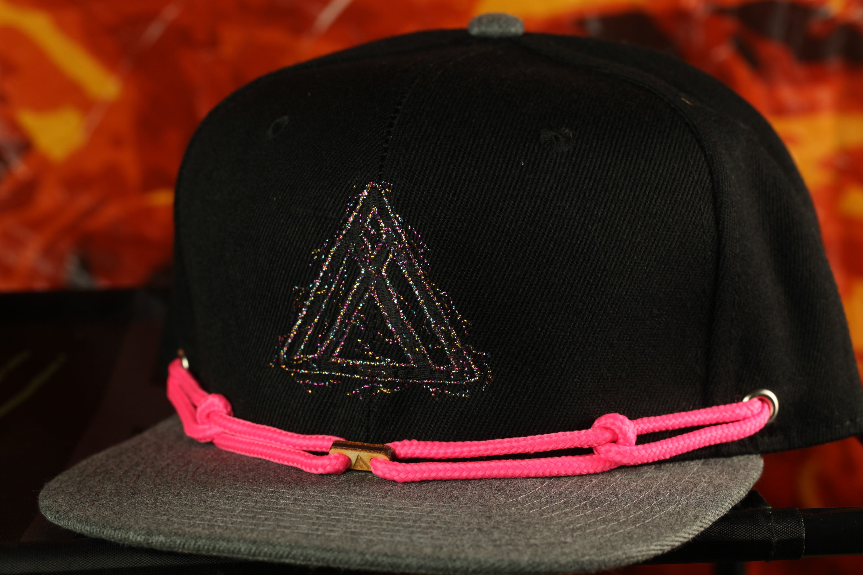 A black baseball cap with a pink braided strap is displayed against a fiery red background.