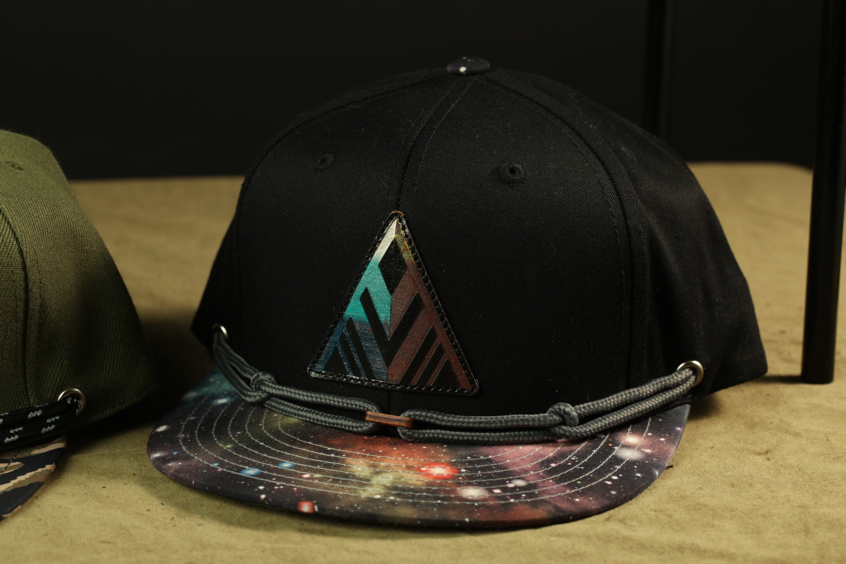 A black baseball cap with a colorful, abstract design on the front, sitting on a surface with a beige background.