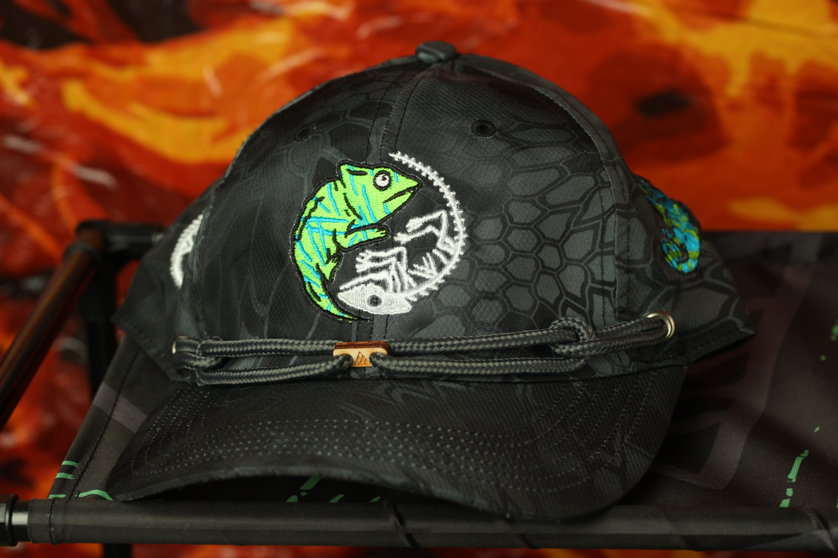 A black baseball cap with a green chameleon design and a geometric pattern on the front.
