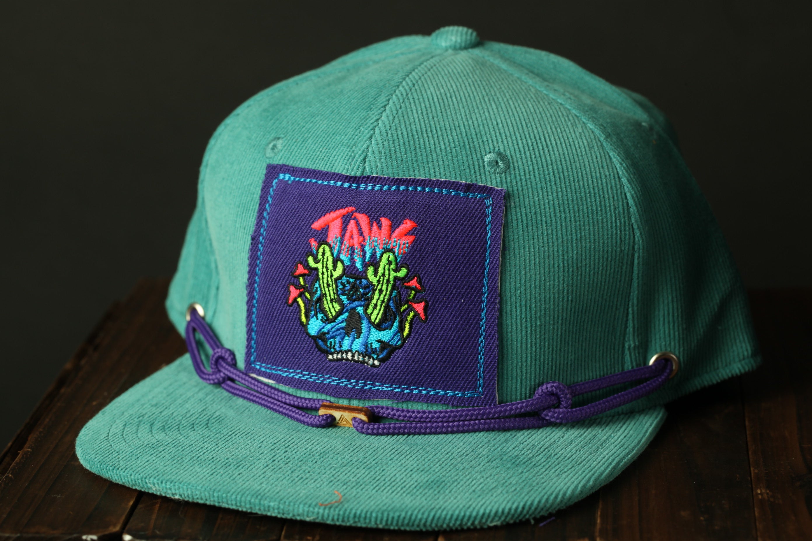 A turquoise baseball cap with a purple band and a patch featuring a graphic design.