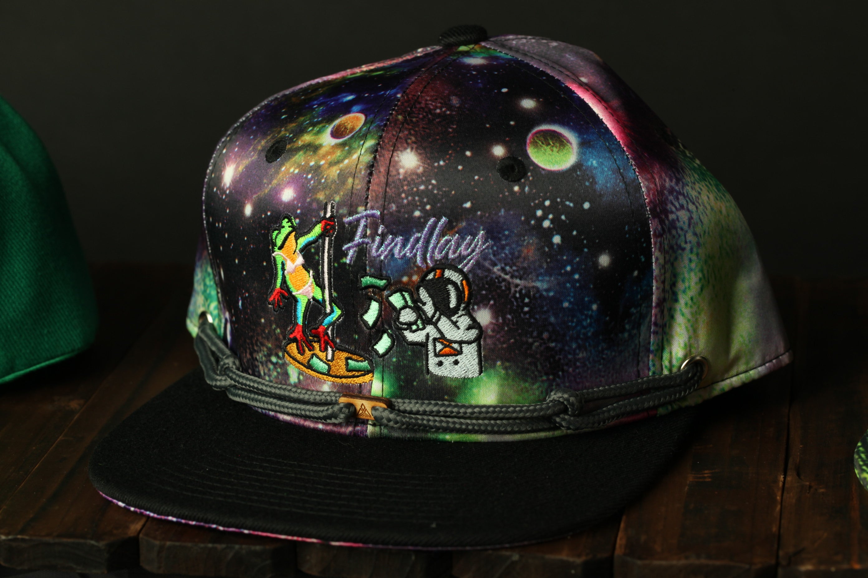 A colorful, space-themed baseball cap with a frog character on it, sitting on a wooden surface.