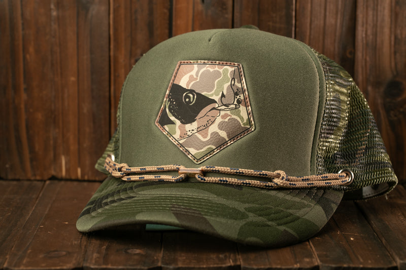A camouflage-patterned baseball cap with a fish patch on the front, resting on a wooden surface.
