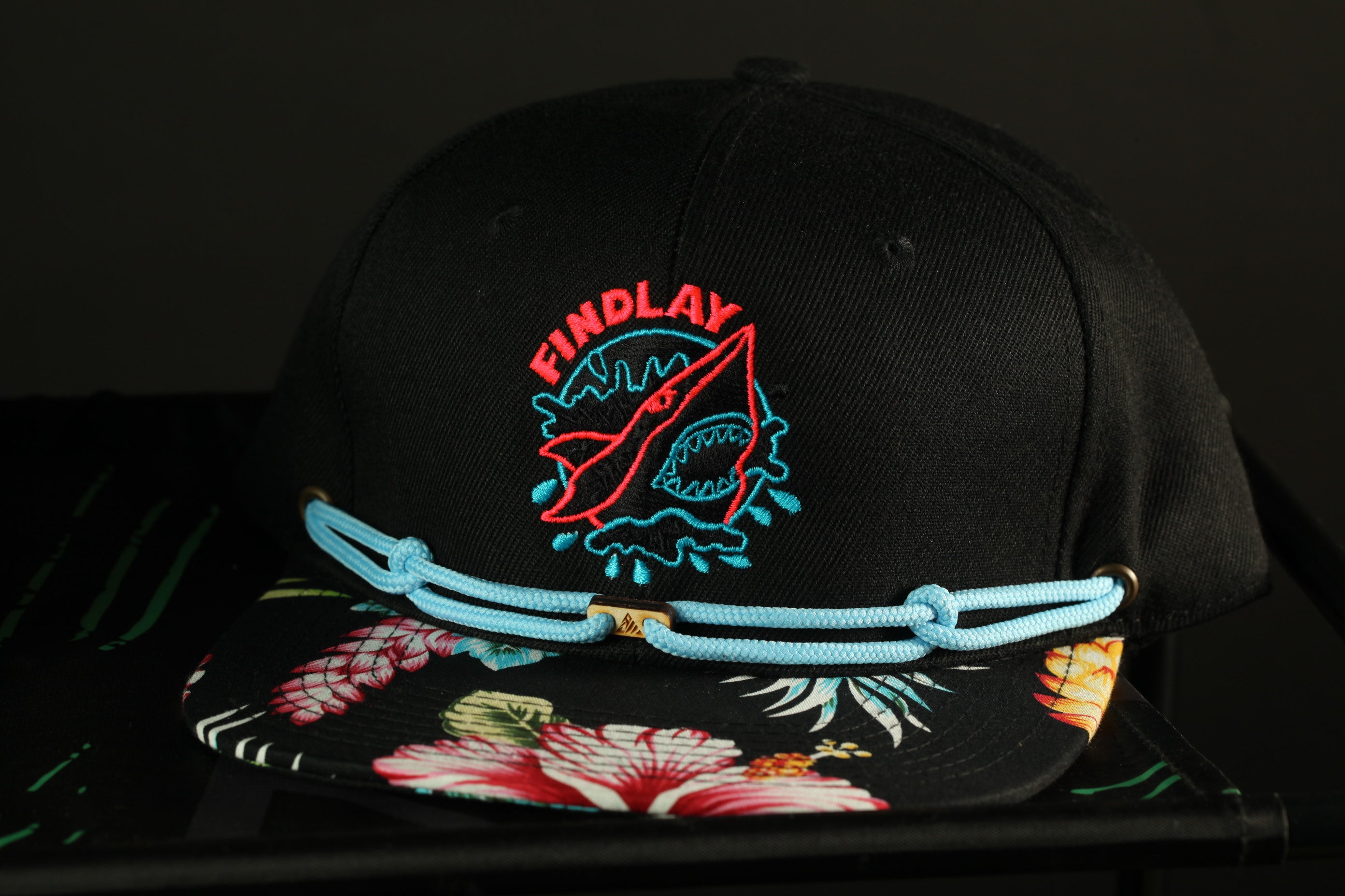 A black baseball cap with a floral pattern and a red and blue logo on the front.
