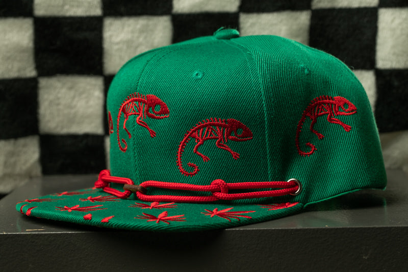 A green baseball cap with red chameleons printed on it, sitting on a black surface against a checkered background.