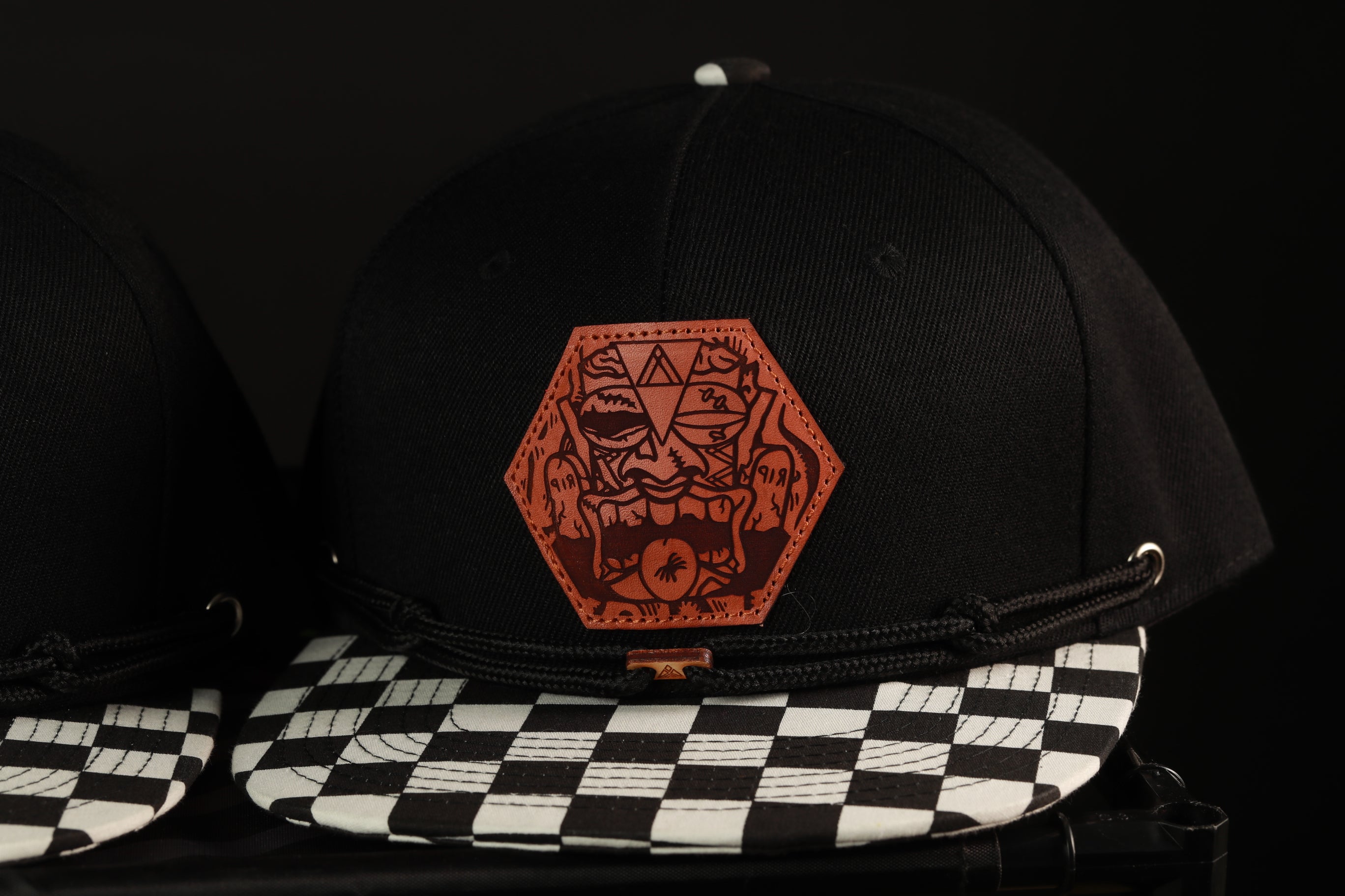A black baseball cap with a checkered pattern and a leather patch featuring an abstract design on the front.
