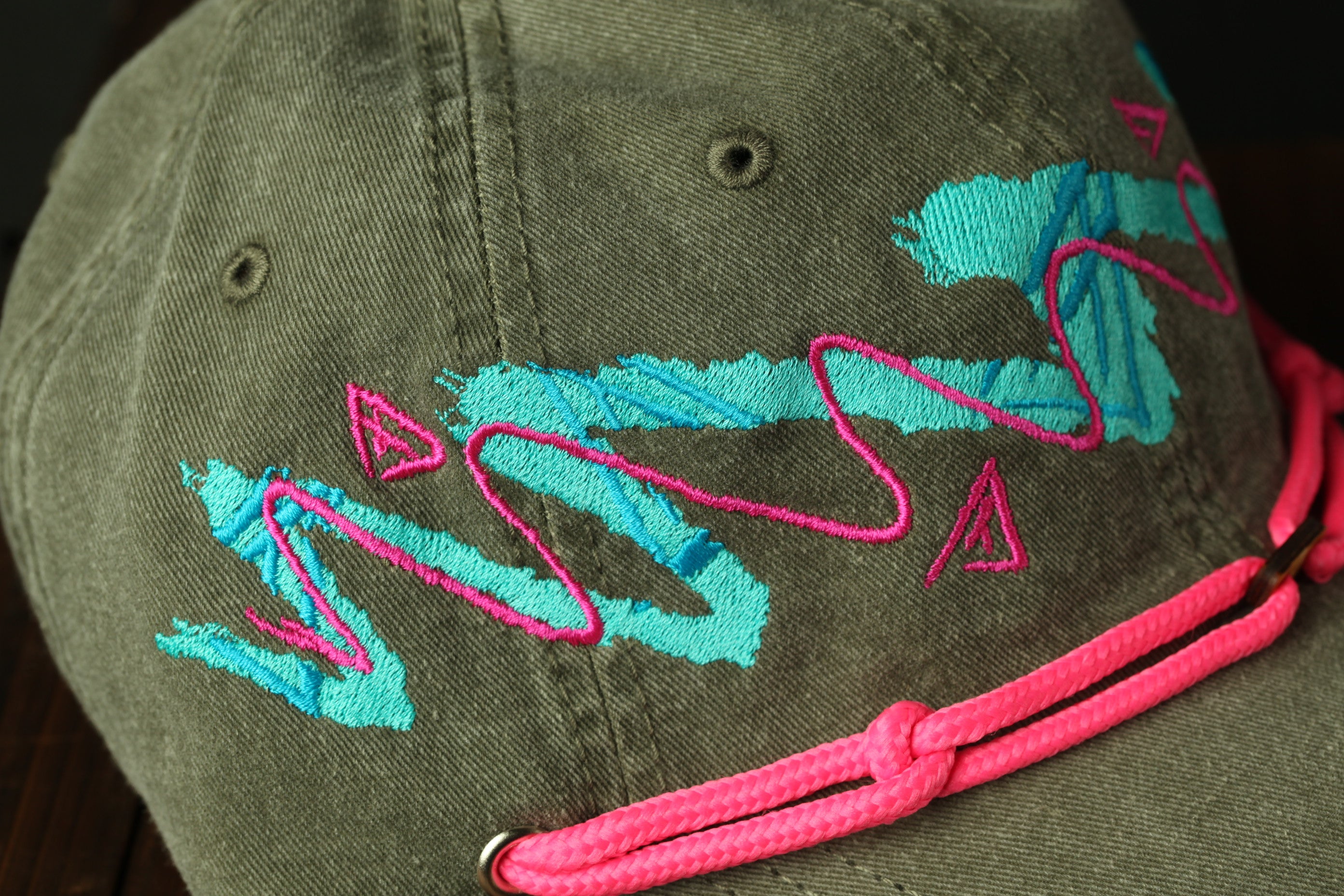 A close-up view of a green baseball cap with a pink braided strap and colorful embroidered designs.