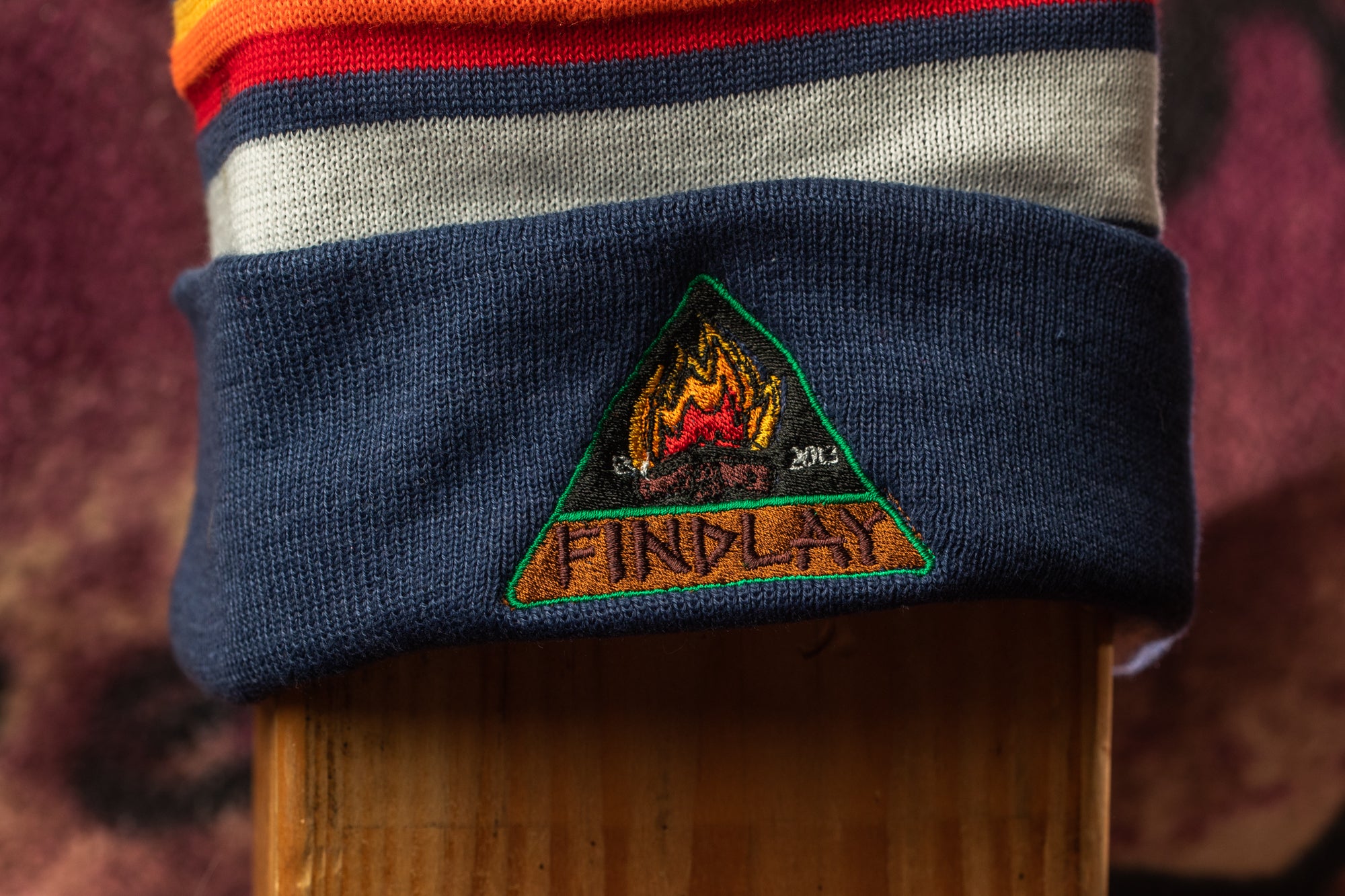 A close-up image of a striped beanie with a logo on it, featuring a triangle with a flame design and the word "FINPLAY" written inside.