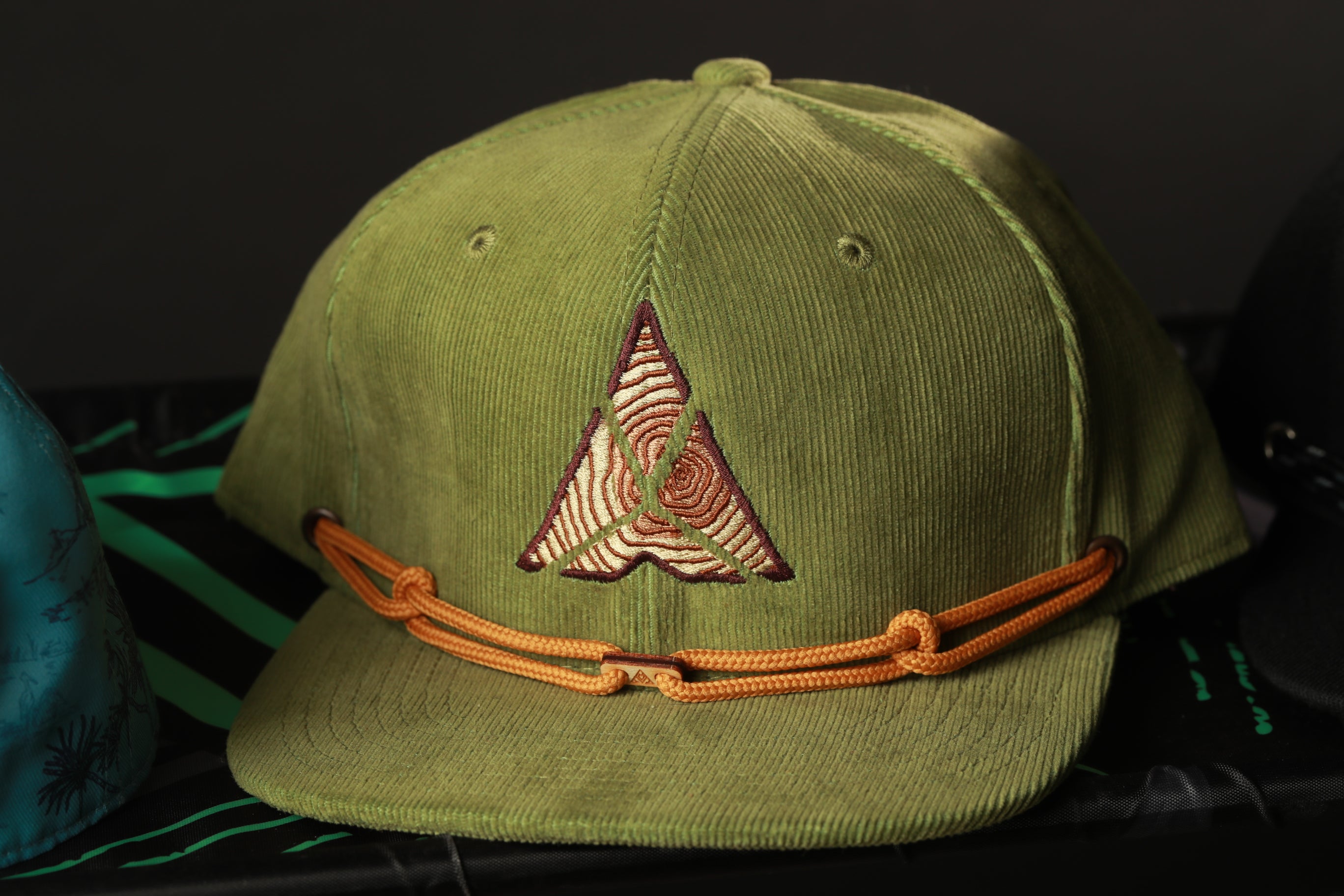 A green baseball cap with an orange adjustable strap and a unique triangular embroidered logo on the front.
